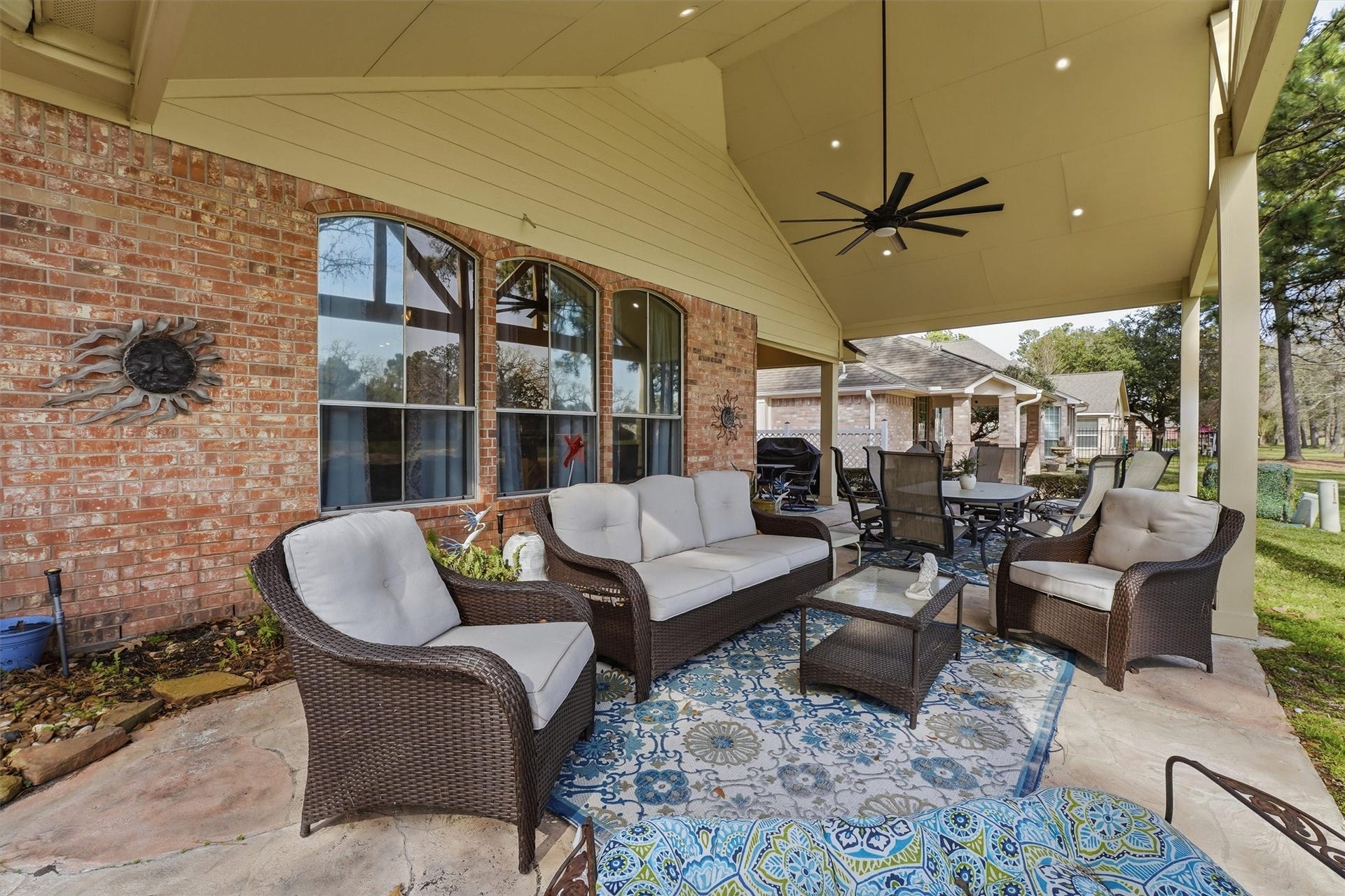 This photo showcases a spacious, covered patio with comfortable wicker furniture and a ceiling fan, set against a brick exterior. The area is perfect for outdoor entertaining, featuring a decorative rug and a peaceful garden view.