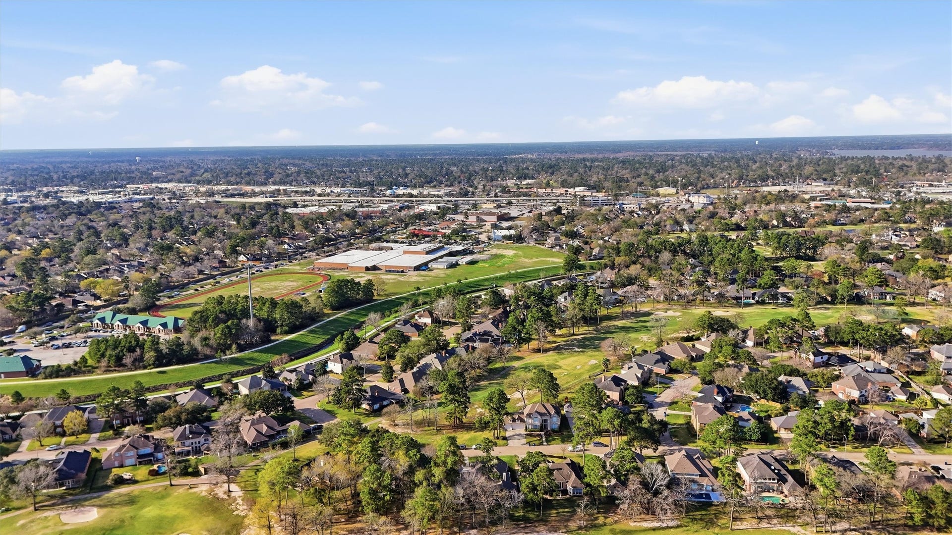 This aerial photo showcases a suburban neighborhood with tree-lined streets and spacious homes, near a golf course and recreational fields. The area is surrounded by greenery, providing a peaceful, spacious environment ideal for families.