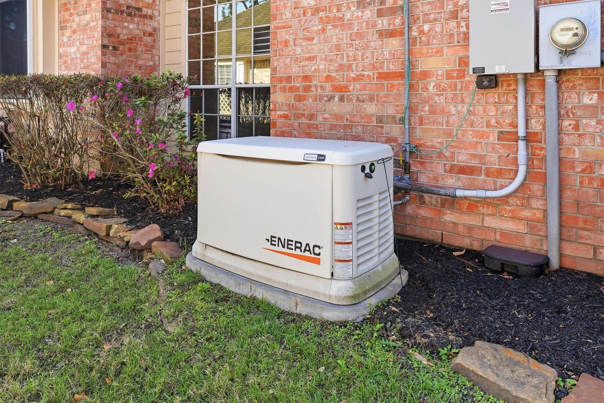 This photo shows an outdoor backup 22K Generac generator next to a brick house, providing reliable power during outages. It's surrounded by a neatly maintained garden with blooming shrubs.