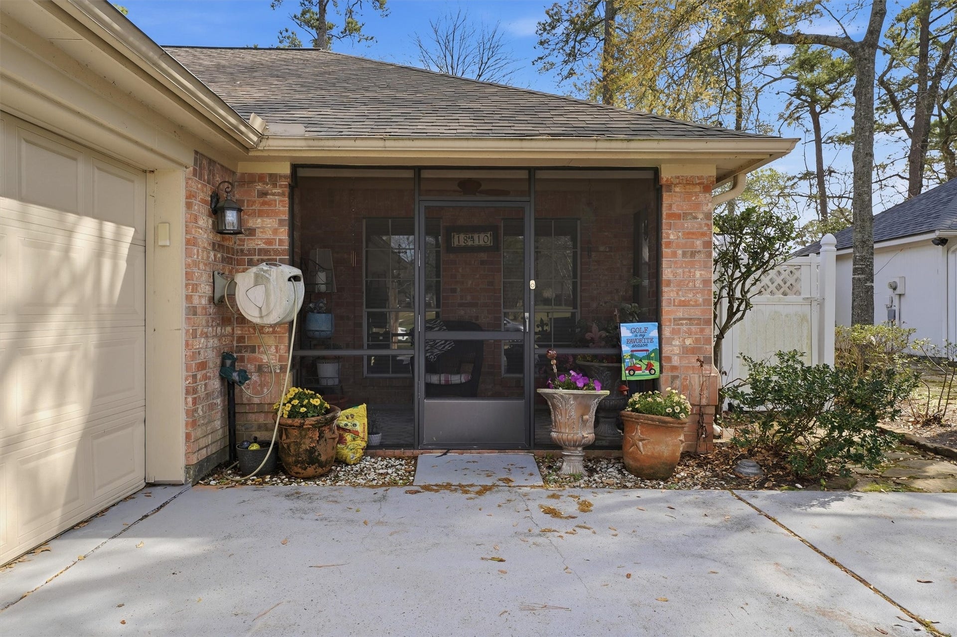 This photo shows a cozy screened-in porch with brick walls, adjacent to a garage. It features potted plants, a hose reel, and a welcoming outdoor seating area, set against a backdrop of trees and a fenced yard.