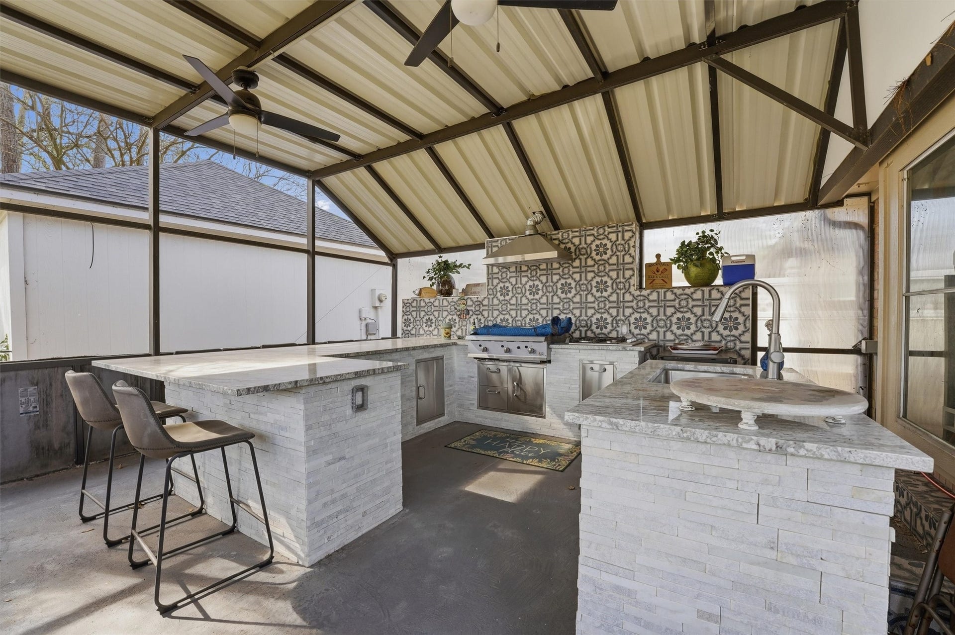 This photo showcases a stylish outdoor kitchen with a covered patio. It features a modern design with a built-in grill, granite countertops, patterned backsplash, and seating at the bar. Ceiling fans provide ventilation, making it perfect for entertaining.