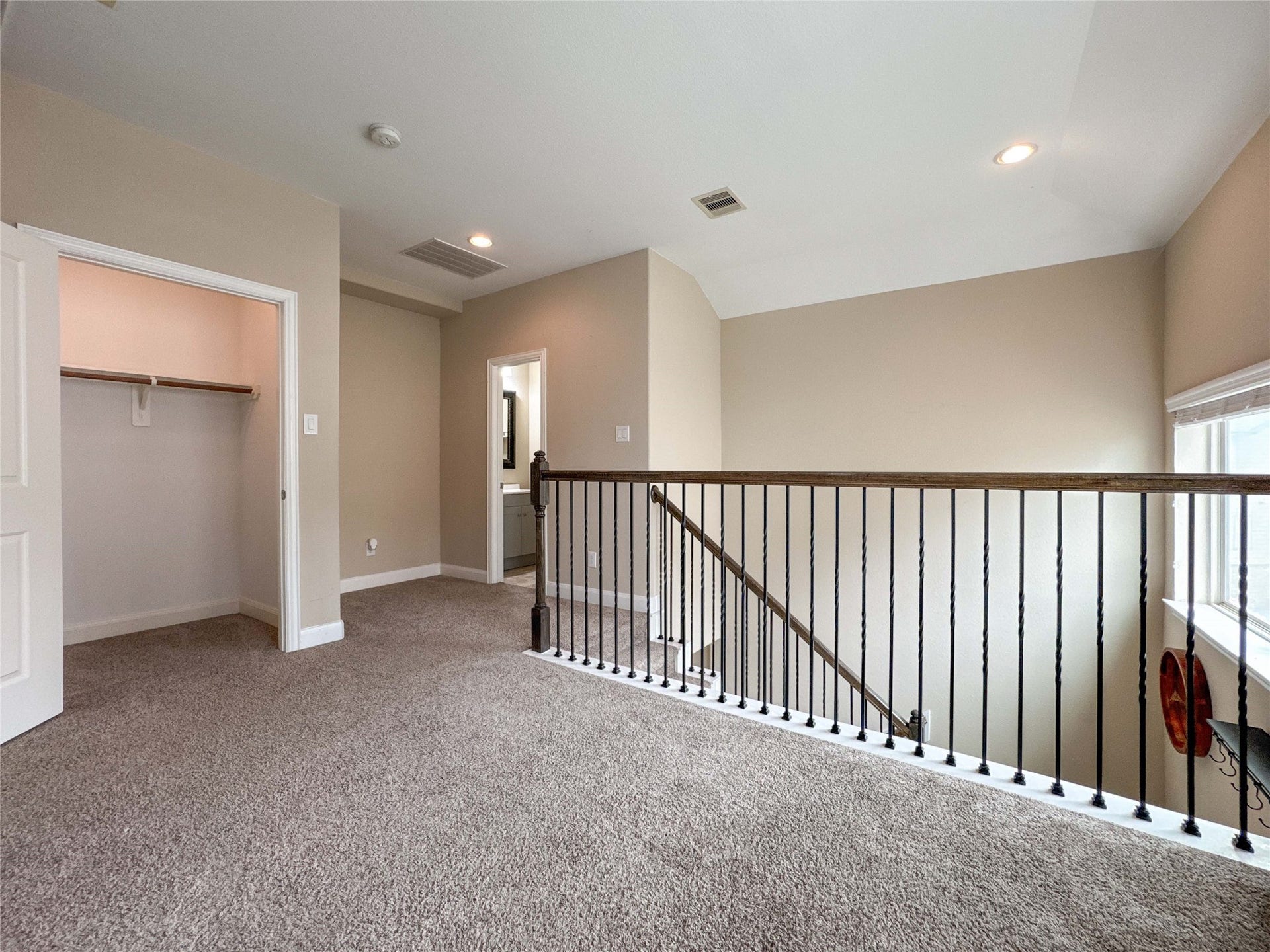 This photo shows a carpeted upstairs landing area with neutral walls and recessed lighting. It features an open railing overlooking the staircase, a window for natural light, and a walk-in closet. There's also an entrance to what appears to be a bathroom.