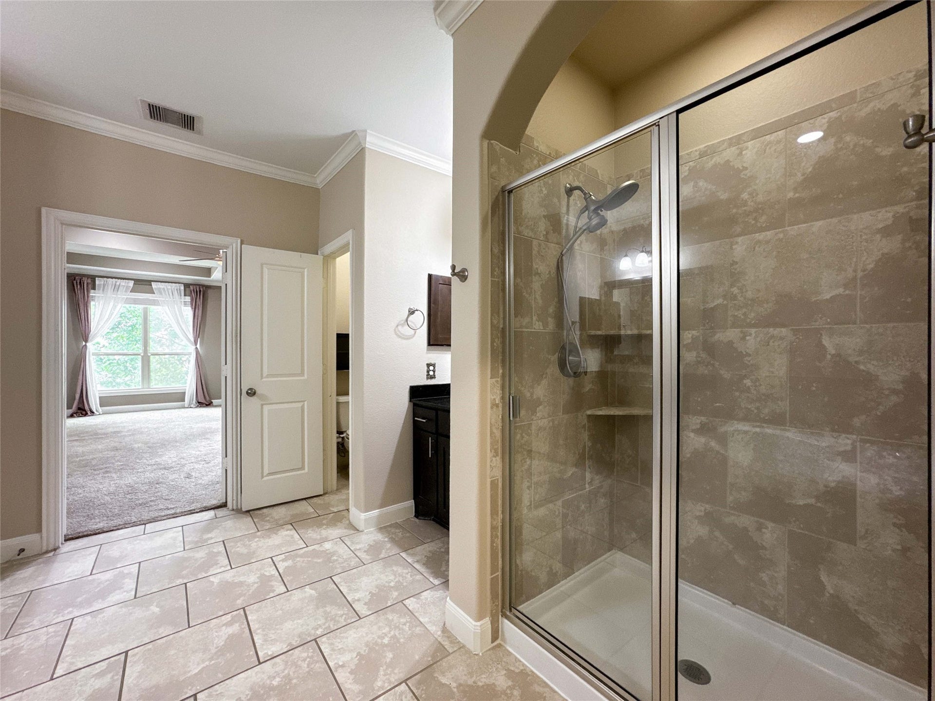 This photo showcases a modern bathroom with a spacious glass-enclosed shower featuring tiled walls. The room has neutral tones, a large window allowing natural light, and connects to a carpeted room, creating an elegant and open feel.