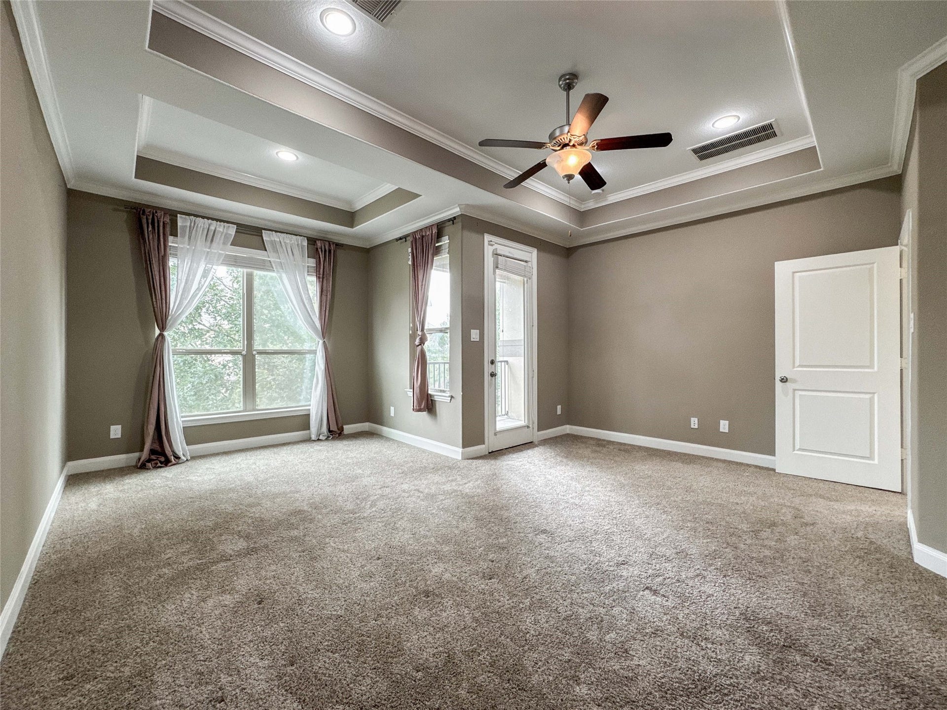 This spacious room features a tray ceiling with recessed lighting, a ceiling fan, neutral carpet, and large windows with elegant drapes, providing ample natural light. A door leads to an outdoor area, and the overall design is modern and inviting.