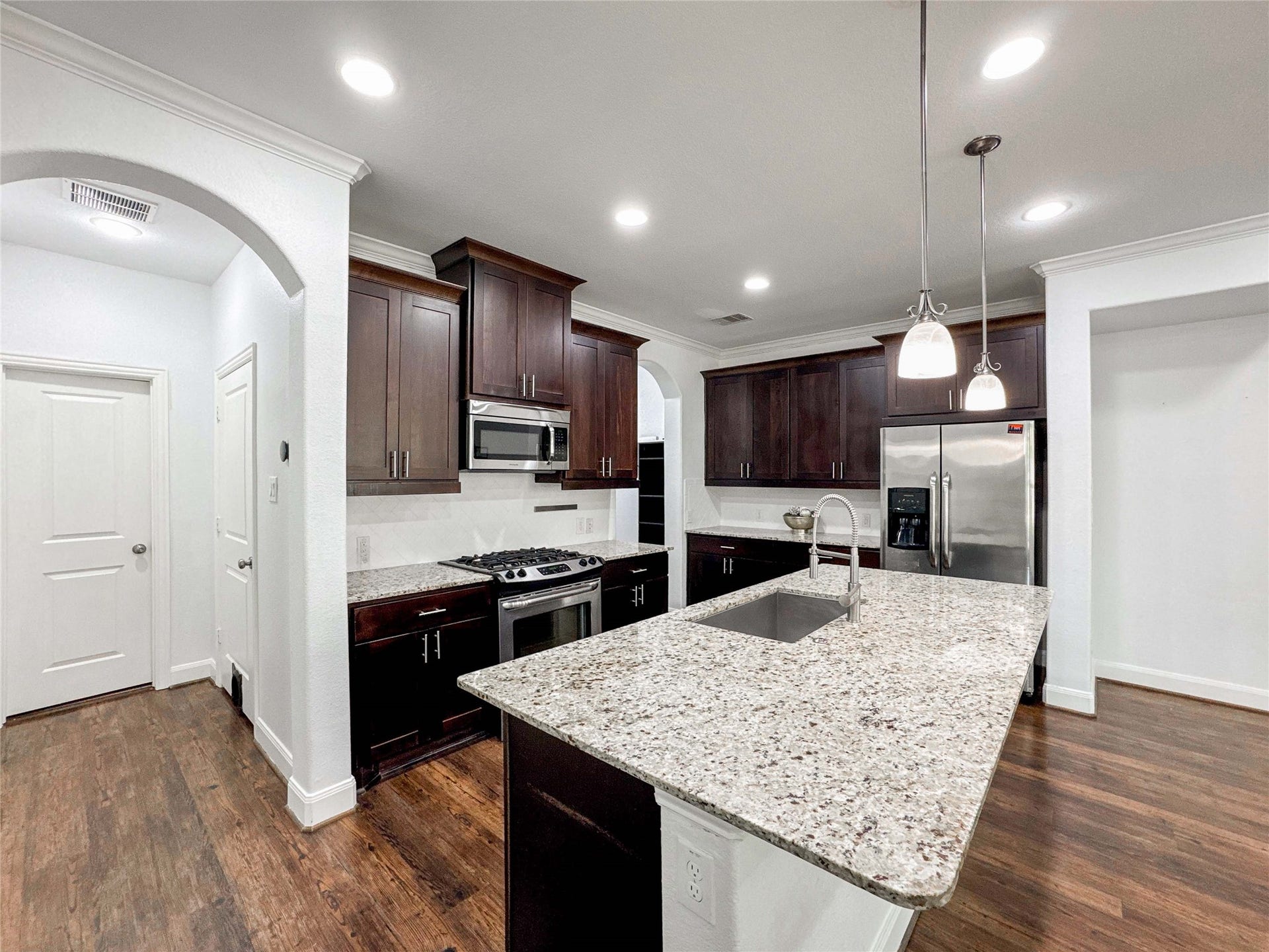 Modern kitchen with dark wood cabinets, granite countertops, and stainless steel appliances. Features an island with a sink and pendant lighting, alongside wood flooring and white walls for a clean, inviting look.