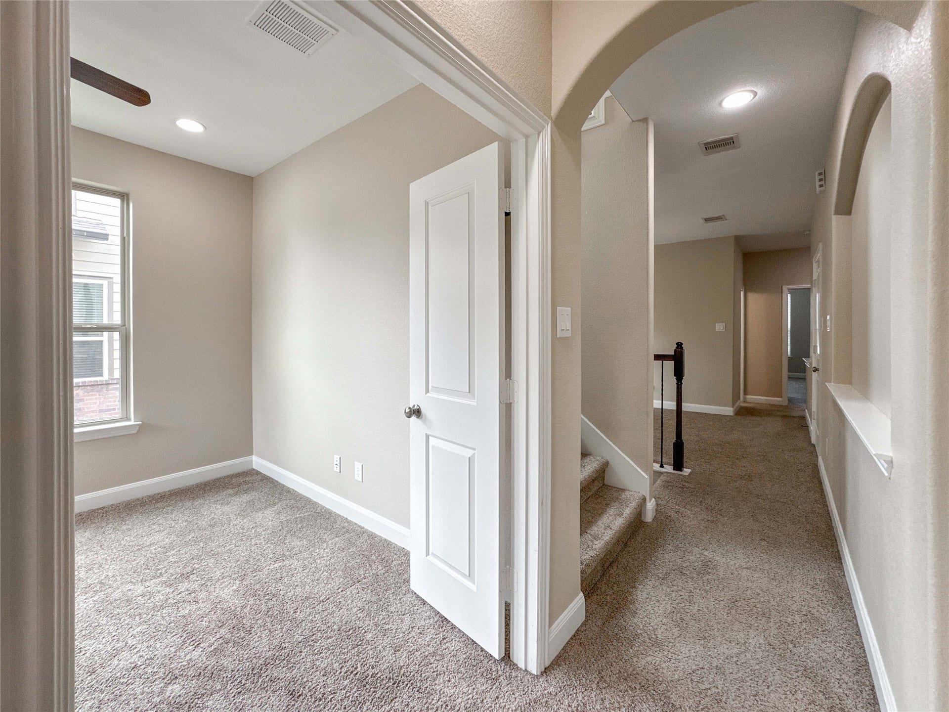 This photo showcases a cozy carpeted hallway with an adjacent room featuring natural light from a window. The hallway leads to a staircase and additional rooms, creating a warm and inviting environment suitable for a comfortable home.