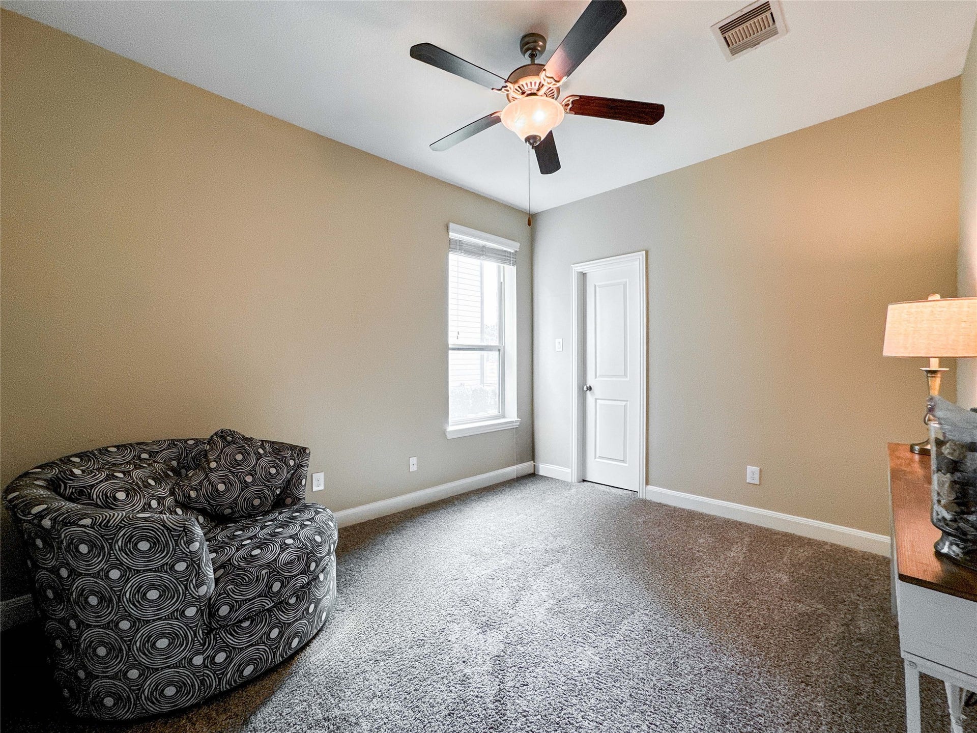 This first floor bedroom features neutral walls and carpet, a ceiling fan with light, and a cozy armchair. It has a window for natural light and a door leading to an adjoining space. Perfect for a small living area or office.