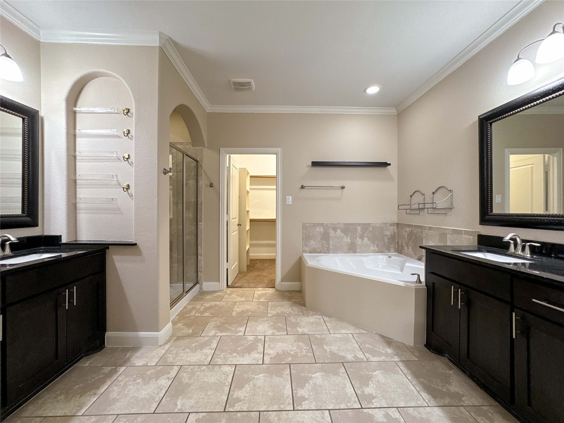 This spacious bathroom features dual vanities with dark wood cabinetry, a large corner bathtub, a glass-enclosed shower, and a walk-in closet, all accented by neutral tiles and soft lighting.