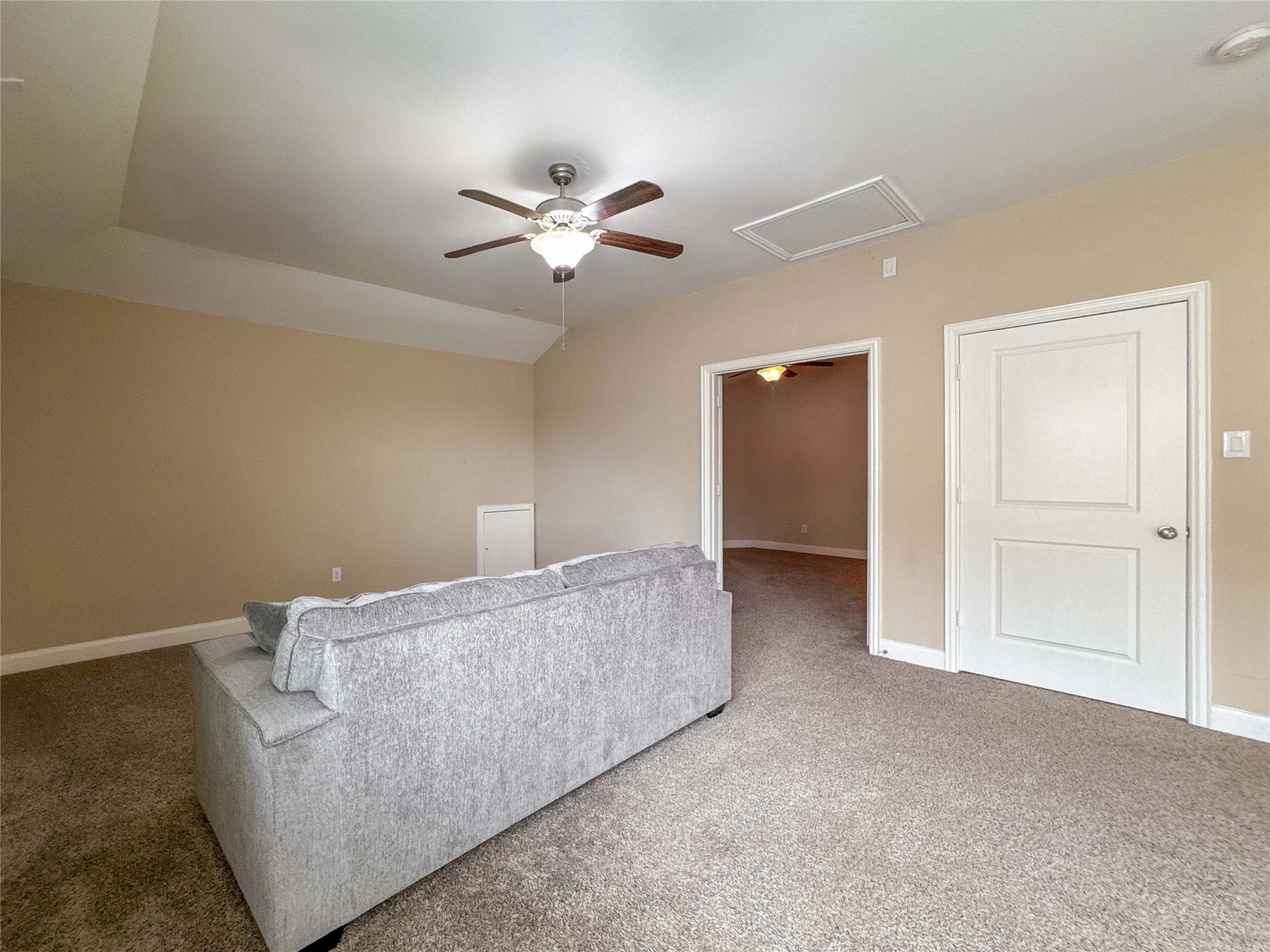 This room features neutral walls and carpeting, a ceiling fan, and a cozy sofa. It leads into another room, providing an open yet defined space.