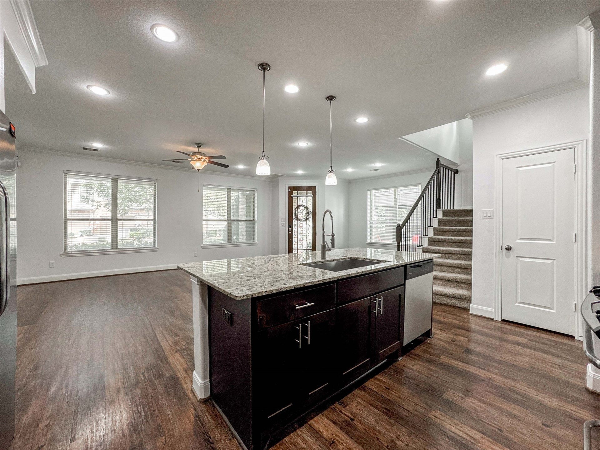This open-concept living space features a modern kitchen with a granite island and dark cabinetry, transitioning into a bright living area with large windows and hardwood floors. A staircase leads to the upper level, and recessed lighting enhances the spacious ambiance.