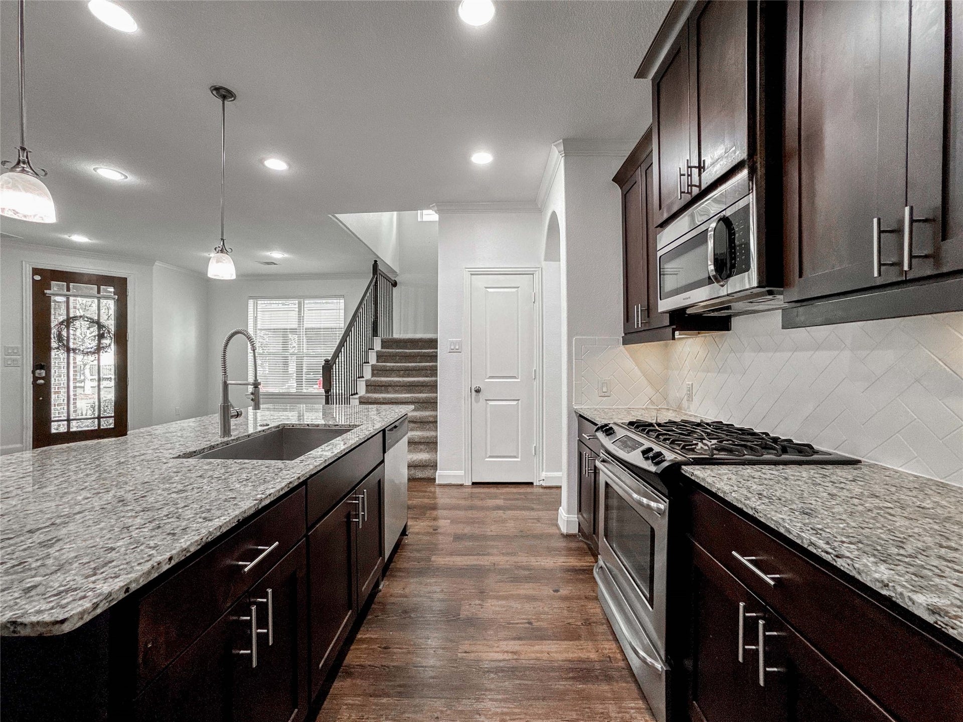 This modern kitchen features sleek dark wood cabinets, granite countertops, and stainless steel appliances. A large island with a sink provides ample prep space. The open layout leads to a bright living area with a staircase, enhancing the home's contemporary appeal.