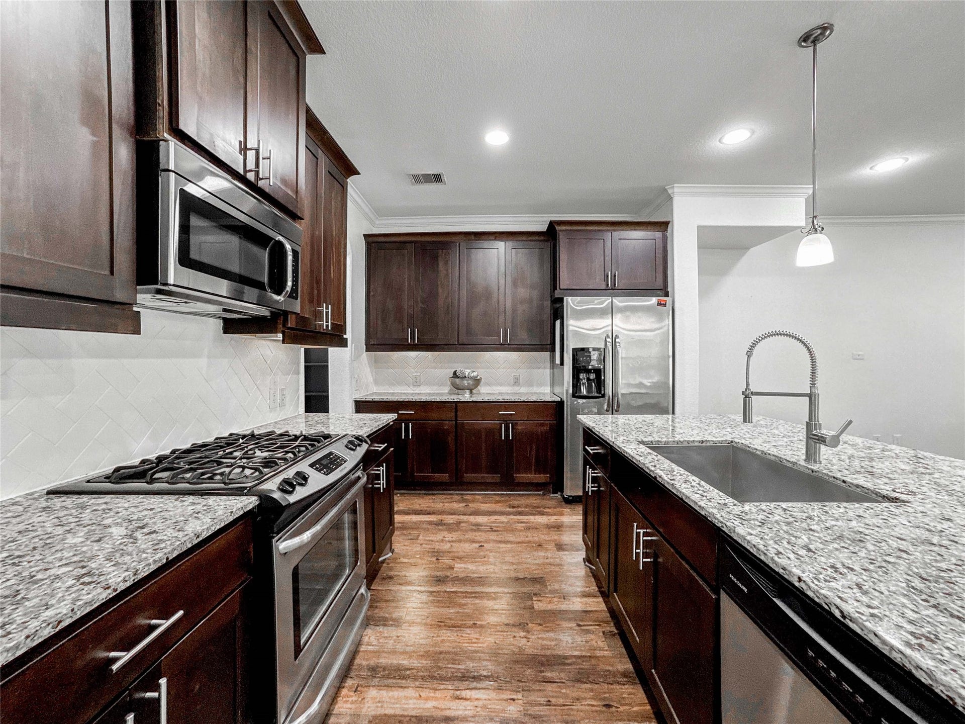 This modern kitchen features rich dark wood cabinets, granite countertops, and stainless steel appliances. It includes a spacious island with a built-in sink, a 4 burner gas stove, and ample storage, creating an inviting space for cooking and entertaining.