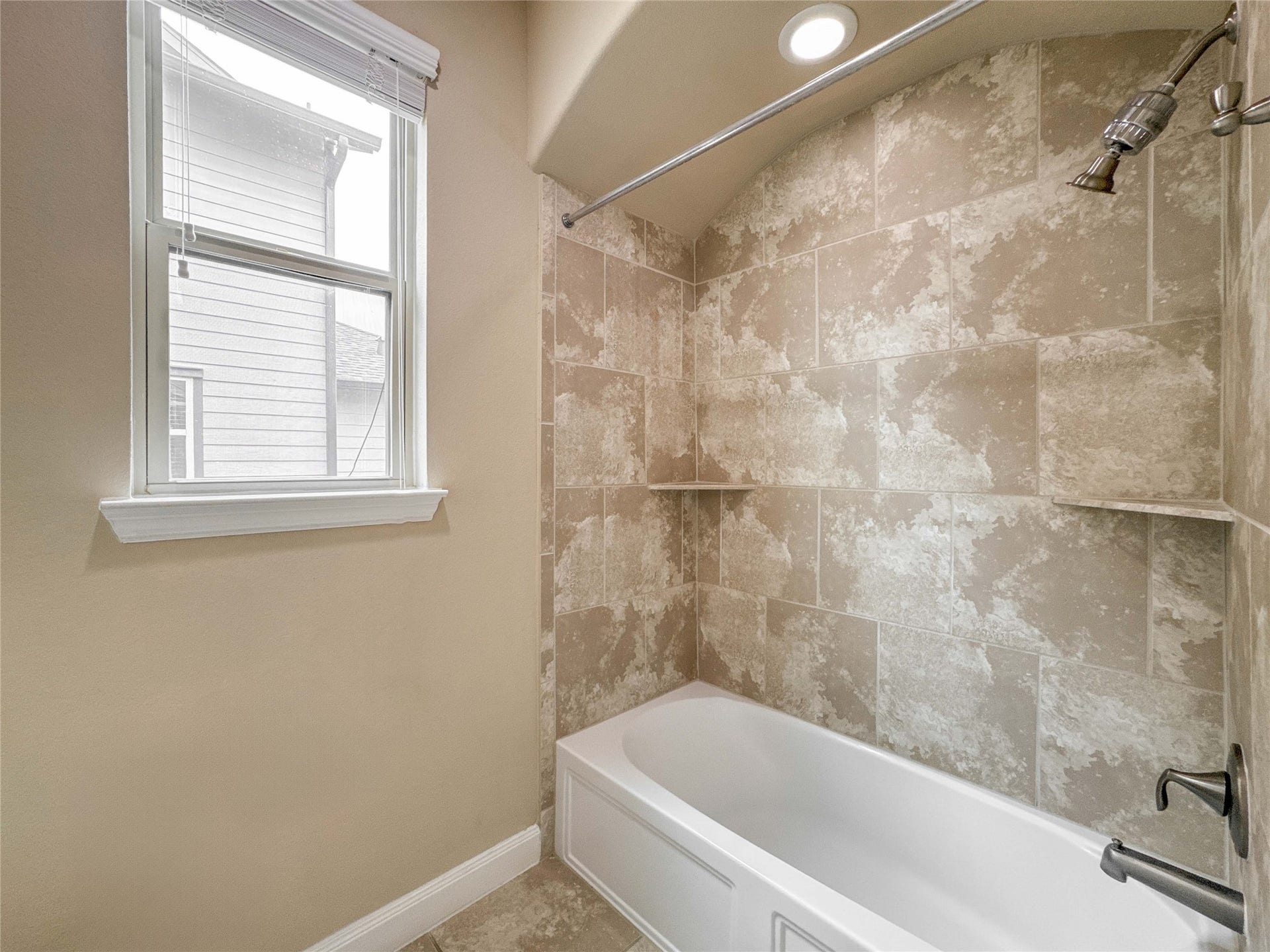 This bathroom features a modern tub and shower combo with elegant beige tile surround, a built-in shelf, and a recessed light above. A window provides natural light, enhancing the airy feel.