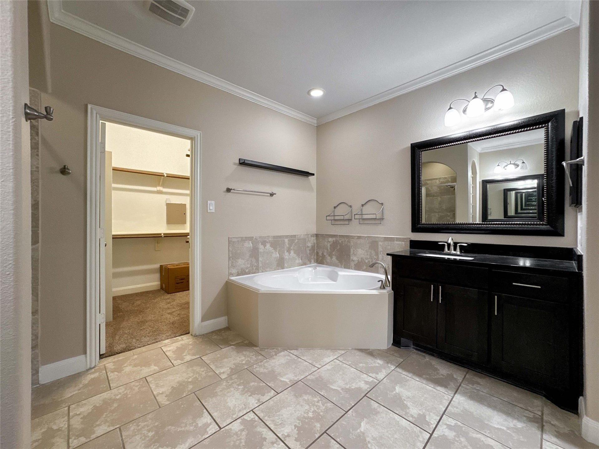 This bathroom features a corner soaking tub, a sleek vanity with a large framed mirror, and tiled flooring. It also includes a spacious walk-in closet for ample storage.