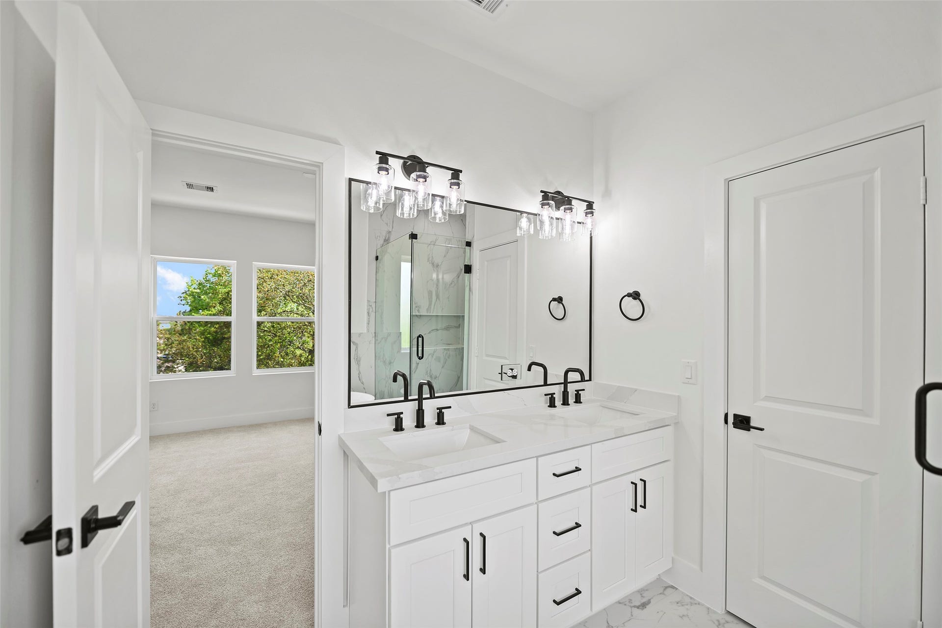 870 W Little York Road, #A, Houston, TX 77091 This bright, modern primary bathroom features a double vanity with sleek black fixtures, elegant lighting, and marble like quartz countertops. It open to the spacious, carpeted primary bedroom with large windows offering a view of lush greenery.