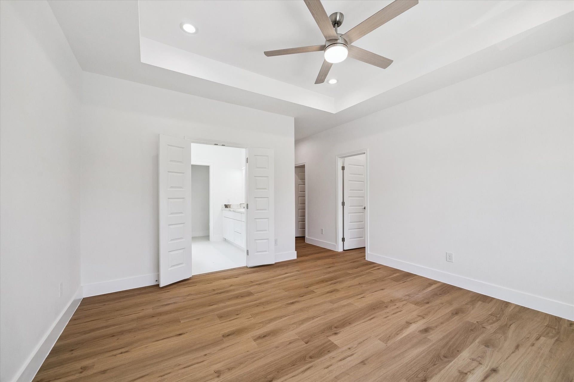 5310 Salina Street, Houston, TX 77026 The spacious primary bedroom offers a clean, modern retreat with warm wood-look flooring, crisp white walls, and a dramatic tray ceiling. A contemporary ceiling fan and recessed lighting add comfort and style, while double doors provide direct access to the adjoining bath. Generous proportions allow plenty of room for a complete bedroom suite, seating area, or additional furnishings.