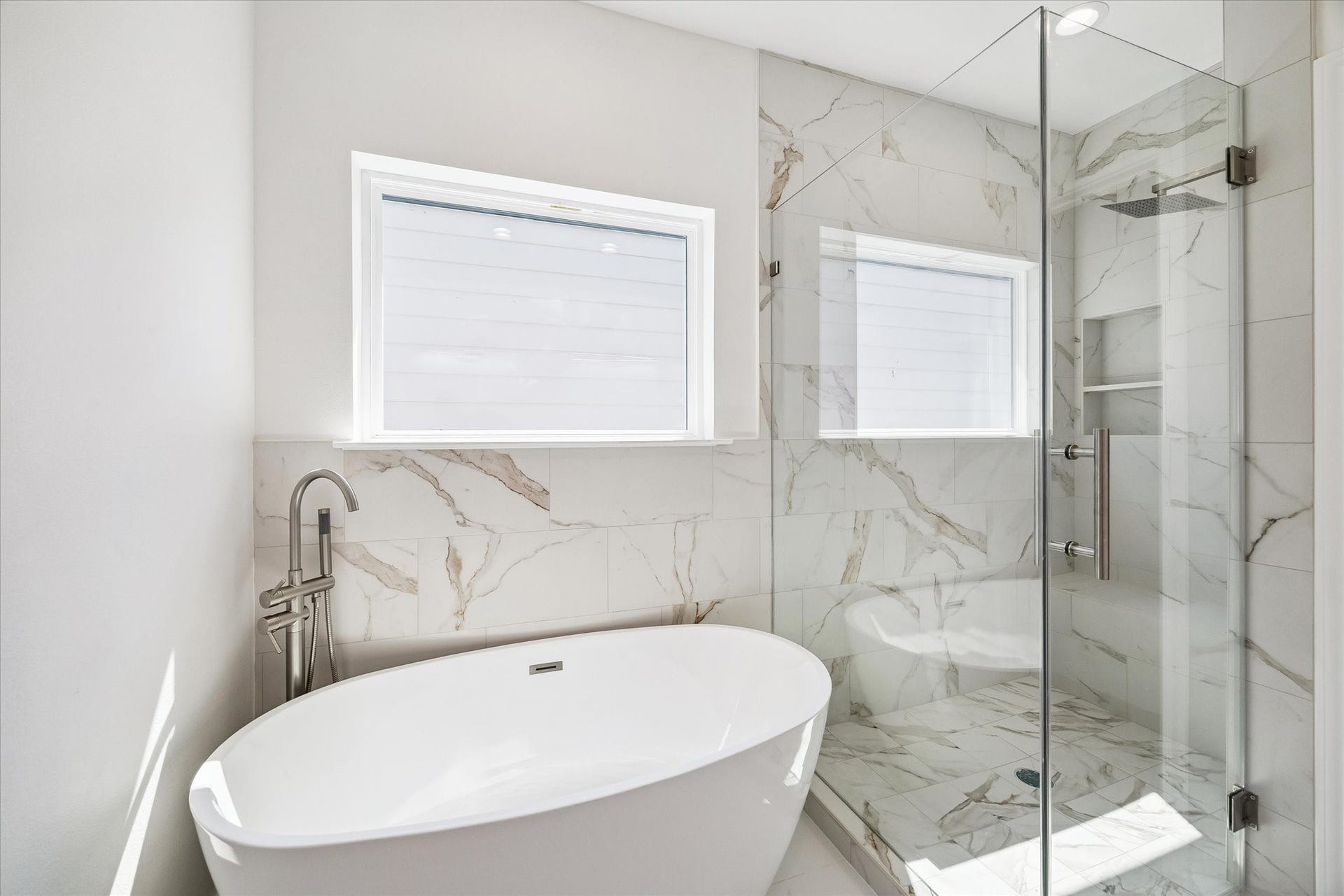 5310 Salina Street, Houston, TX 77026 The primary baths freestanding soaking tub and oversized glass-enclosed shower create a luxurious focal point. Marble-look tile surrounds the bathing area and continues into the shower, where a rainfall-style showerhead and built-in niche enhance the modern design. Natural light from the windows brightens the space, while brushed metal fixtures add a refined finishing touch.