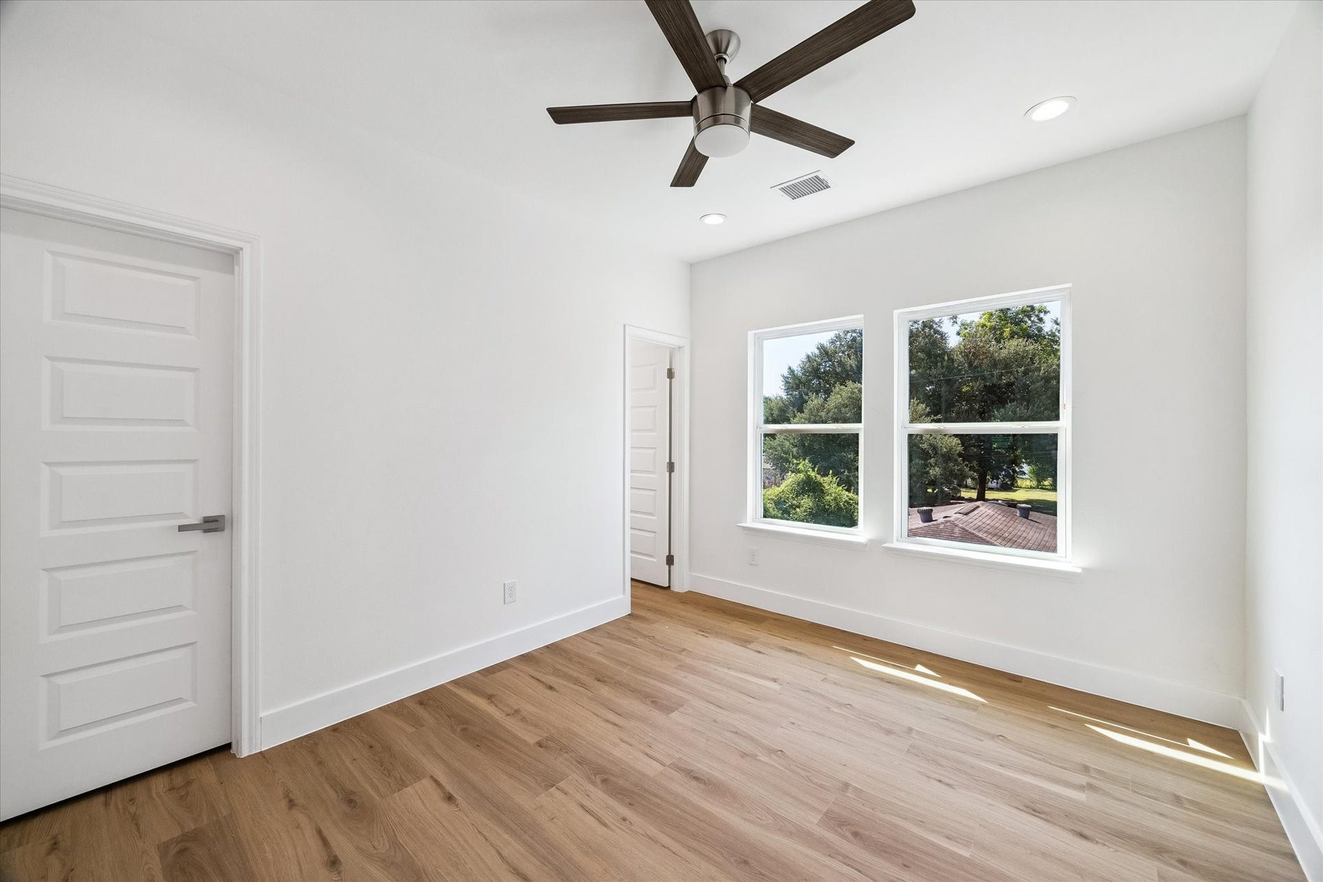 5310 Salina Street, Houston, TX 77026 A spacious secondary bedroom is filled with natural light from two large windows overlooking mature greenery. Warm wood-look flooring, crisp white walls, recessed lighting, and a modern ceiling fan create a bright and comfortable setting. The generous floor plan provides plenty of flexibility for a full bedroom suite, desk, reading area, or additional storage pieces.