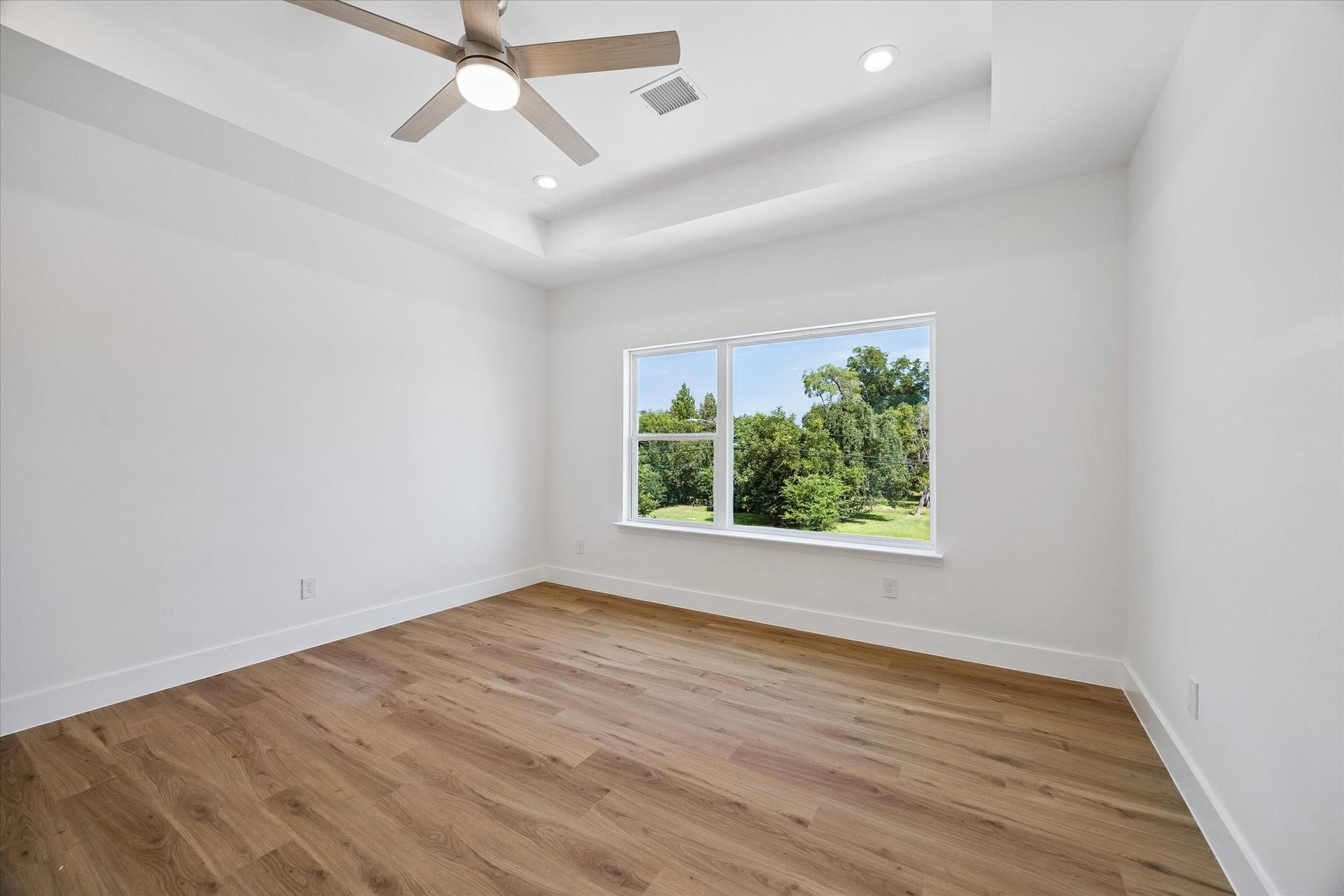 5310 Salina Street, Houston, TX 77026 Another view of the bedroom highlights its generous proportions, elevated tray ceiling, and abundant natural light. The large window frames leafy outdoor views while warm wood-look flooring adds character and continuity. A contemporary ceiling fan and recessed lighting complete the clean, modern presentation. The open floor plan provides flexibility for substantial furniture, a sitting area, or additional storage pieces.