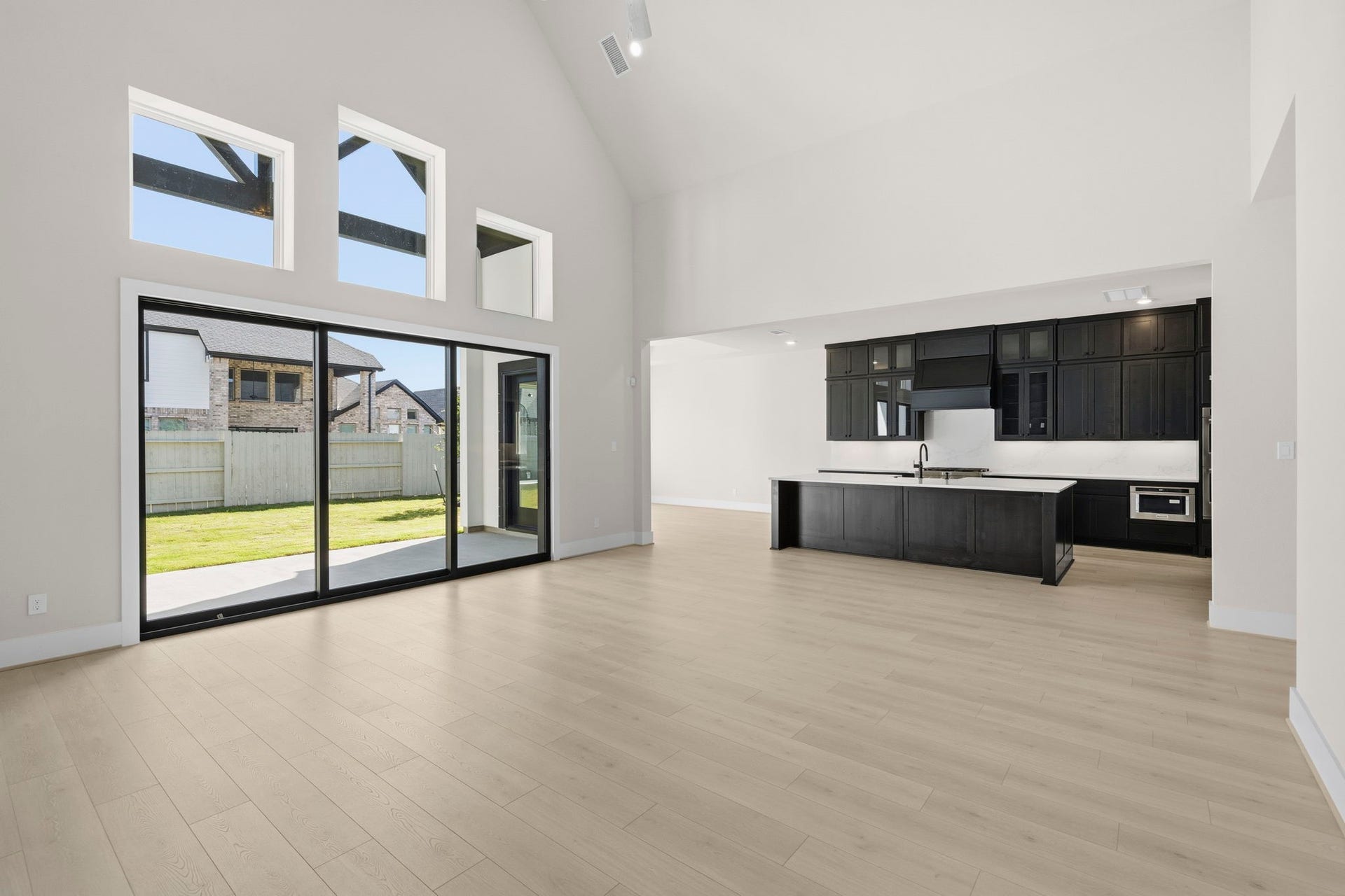 This modern open-concept living space features high ceilings, ample natural light from large windows, and a sleek kitchen with dark cabinetry. The sliding glass doors lead to a spacious backyard, creating an inviting indoor-outdoor flow.