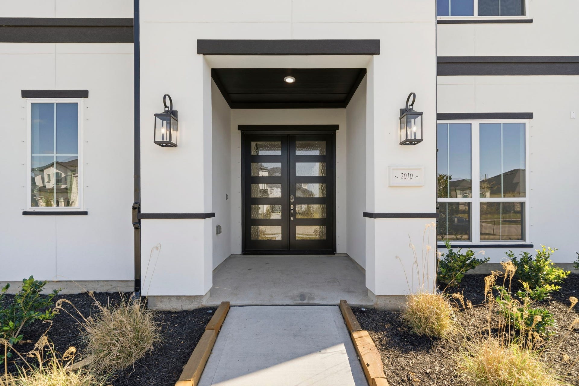 Modern entryway with a black-framed glass door, sleek exterior lighting, and clean landscaping leading up to the entrance.