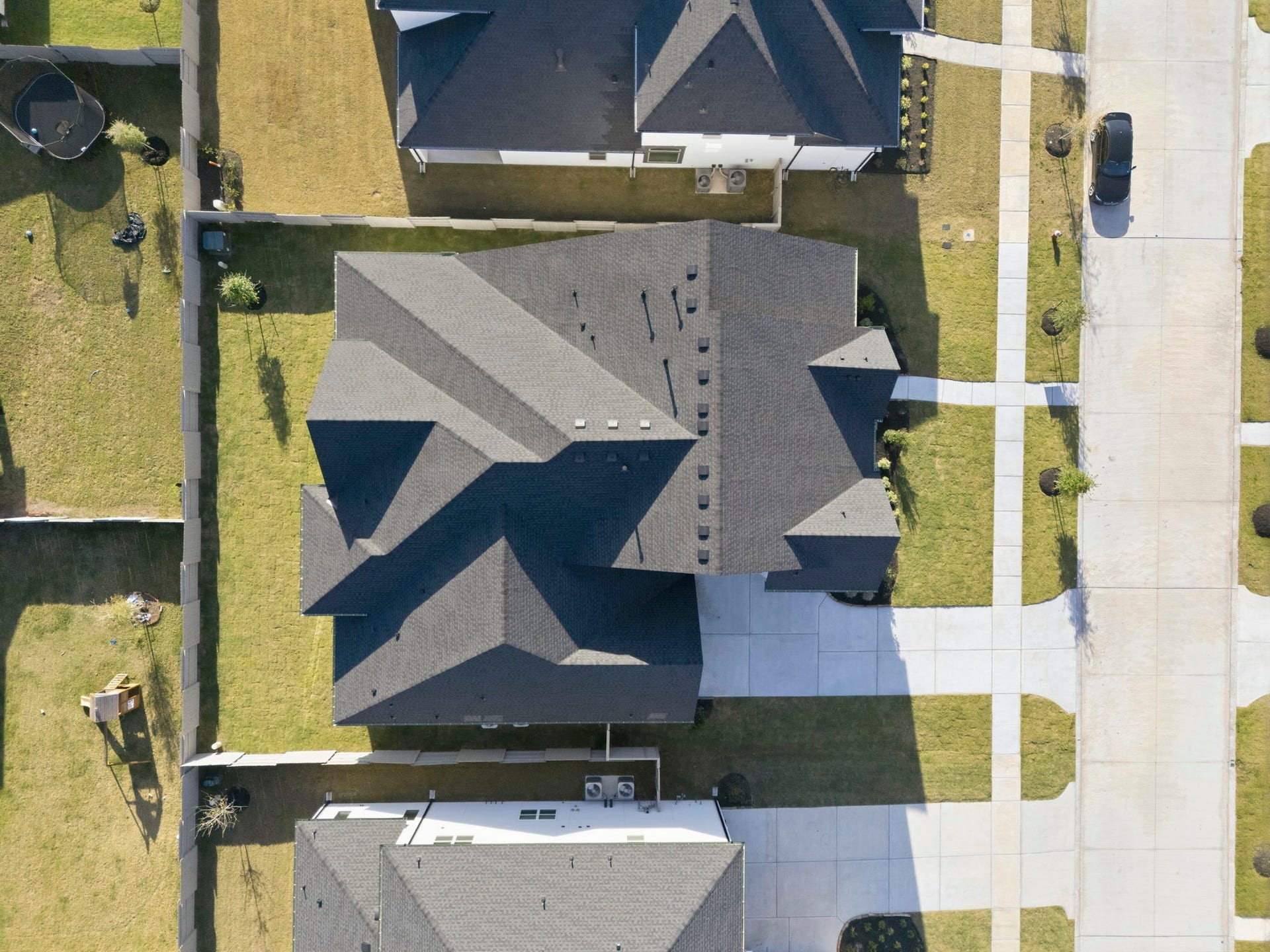 This aerial photo shows a modern, well-maintained home with a dark roof, spacious driveway, and neatly landscaped yard. The property is in a suburban neighborhood with similar houses nearby, offering privacy with fenced boundaries.