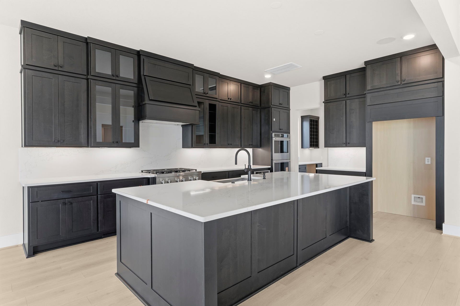 This kitchen features sleek, dark cabinetry with ample storage, a large island with a modern sink, and stainless steel appliances. The light wood flooring and neutral walls create a bright, contemporary space perfect for cooking and entertaining.