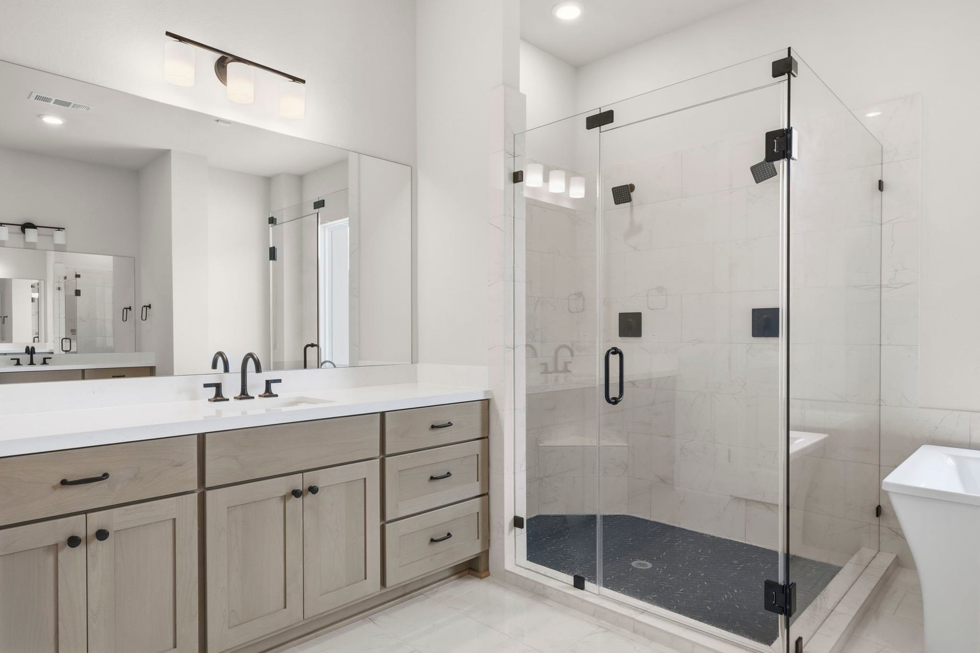 This primary bathroom features a spacious glass-enclosed shower, dual sinks with modern fixtures, light wood cabinetry, and bright lighting, creating a clean, contemporary, and home spa sanctuary