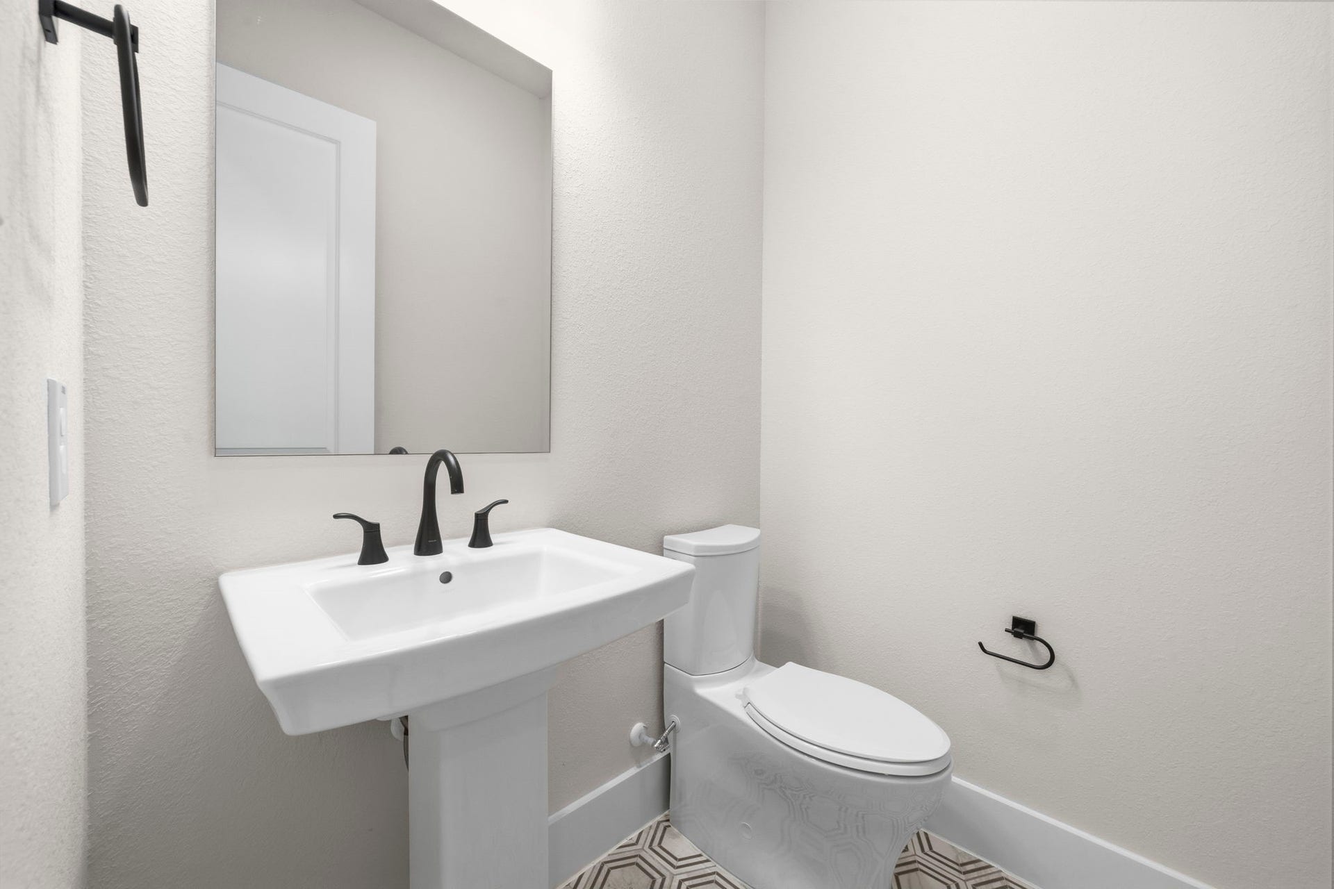 Modern half-bathroom featuring a pedestal sink with sleek black fixtures, a large mirror, and a toilet. The room is bright with neutral walls and patterned floor tiles.