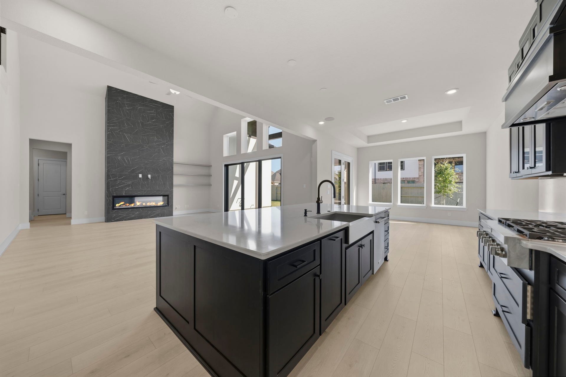 This modern kitchen features sleek dark cabinetry, a large center island with a sink, and stainless steel appliances. The space is brightened by natural light from large windows and has light hardwood flooring.