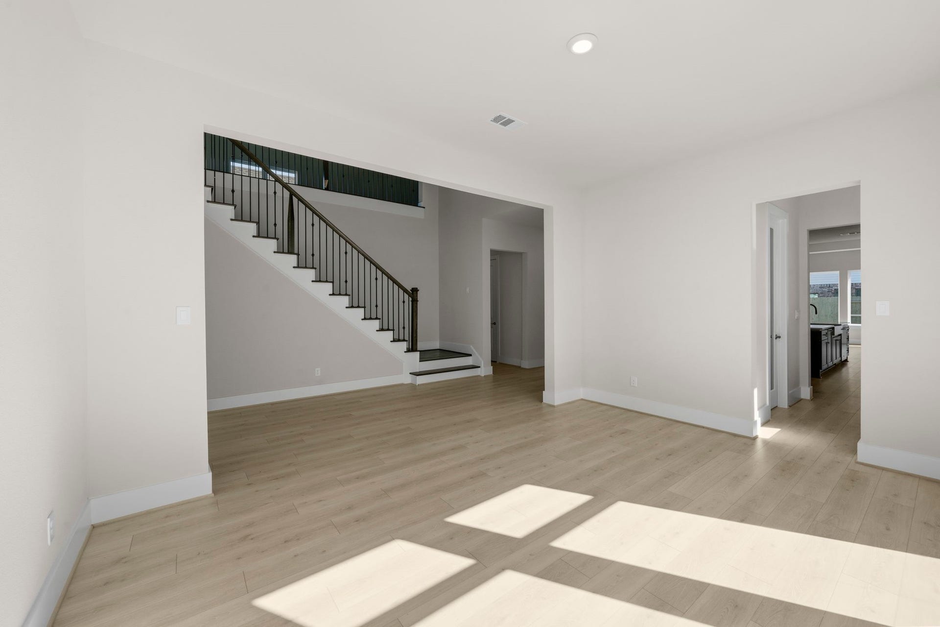 This spacious living area features light wood flooring, neutral walls, and ample natural light. It has an open view of a staircase with elegant railing, and leads into the kitchen, creating a seamless flow.