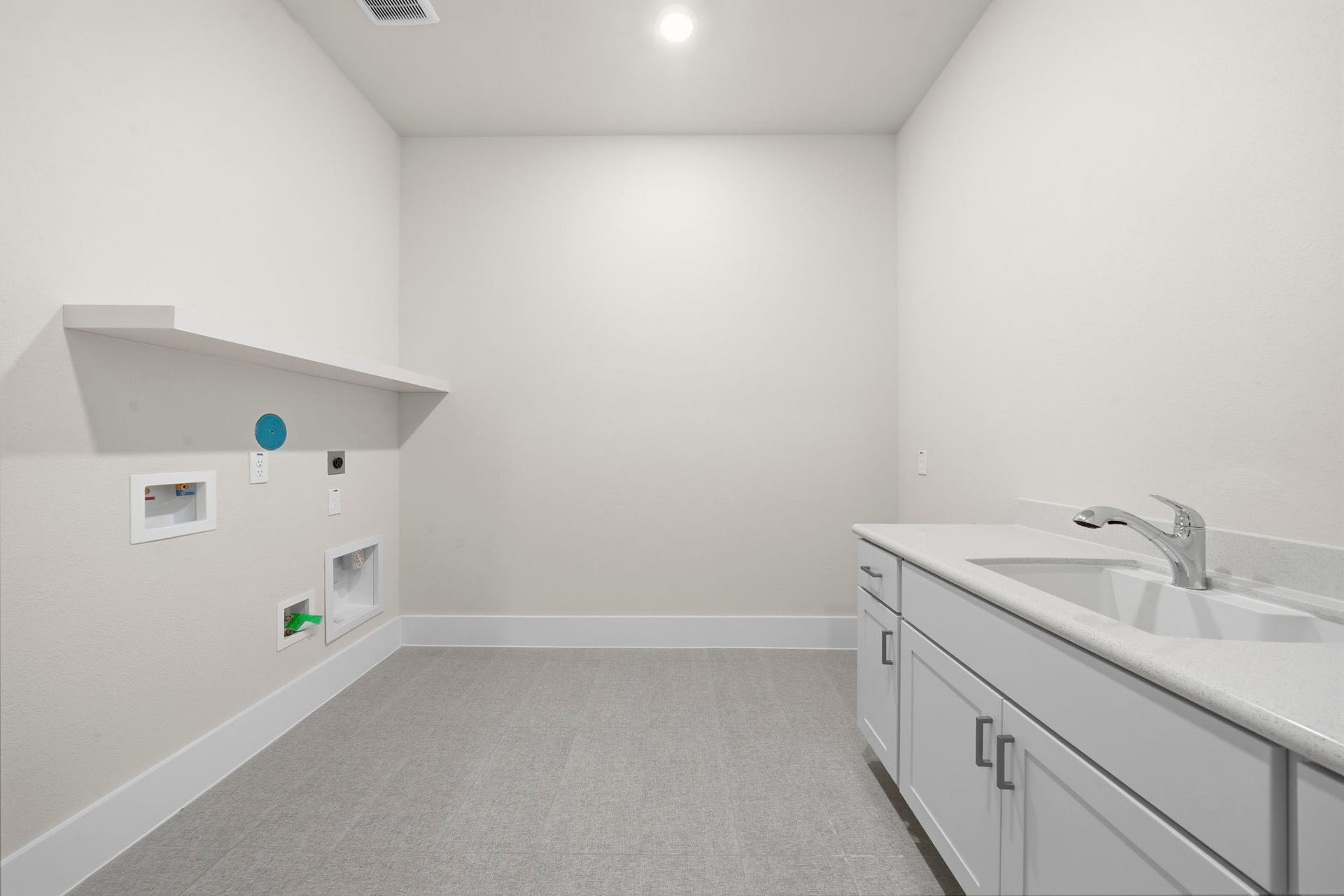 Spacious laundry room with light-colored walls and flooring, featuring a countertop with a sink and ample cabinetry for storage. A shelf and hookups are available for appliances.