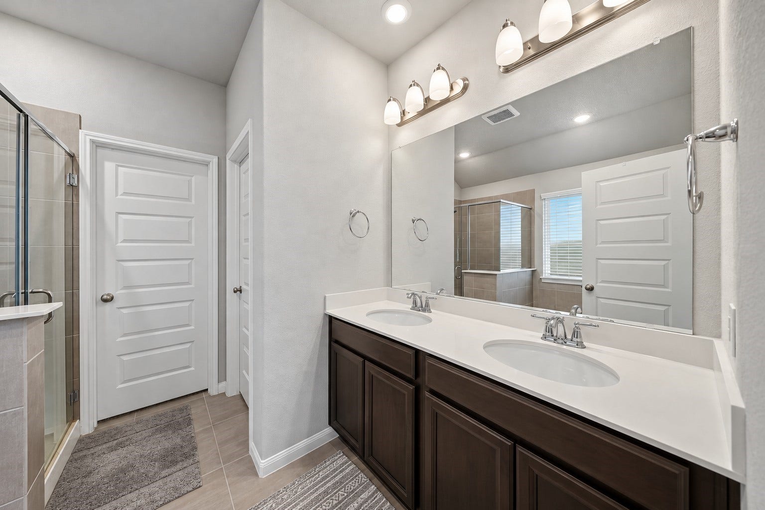 903 Pismo Lane, Rosharon, TX 77583 Virtually Enhanced: This spacious primary bathroom features a double-sink vanity with a sleek white countertop, undermount basins, and rich dark cabinetry offering generous storage. A large wall-to-wall mirror, and recessed ceiling lighting create a bright, welcoming atmosphere. Neutral tile flooring complements the soft gray walls and crisp white trim.