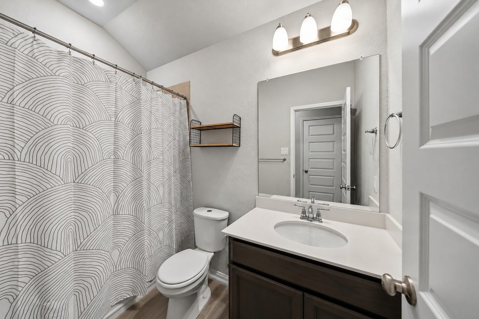 903 Pismo Lane, Rosharon, TX 77583 Virtually Enhanced: This well-appointed bathroom features a clean, functional layout with soft neutral walls and durable wood-look flooring. A spacious single-sink vanity offers a white countertop, an undermount basin, and dark cabinetry for convenient storage. A large wall-mounted mirror, three-light vanity fixture, and recessed ceiling lighting provide excellent illumination.
