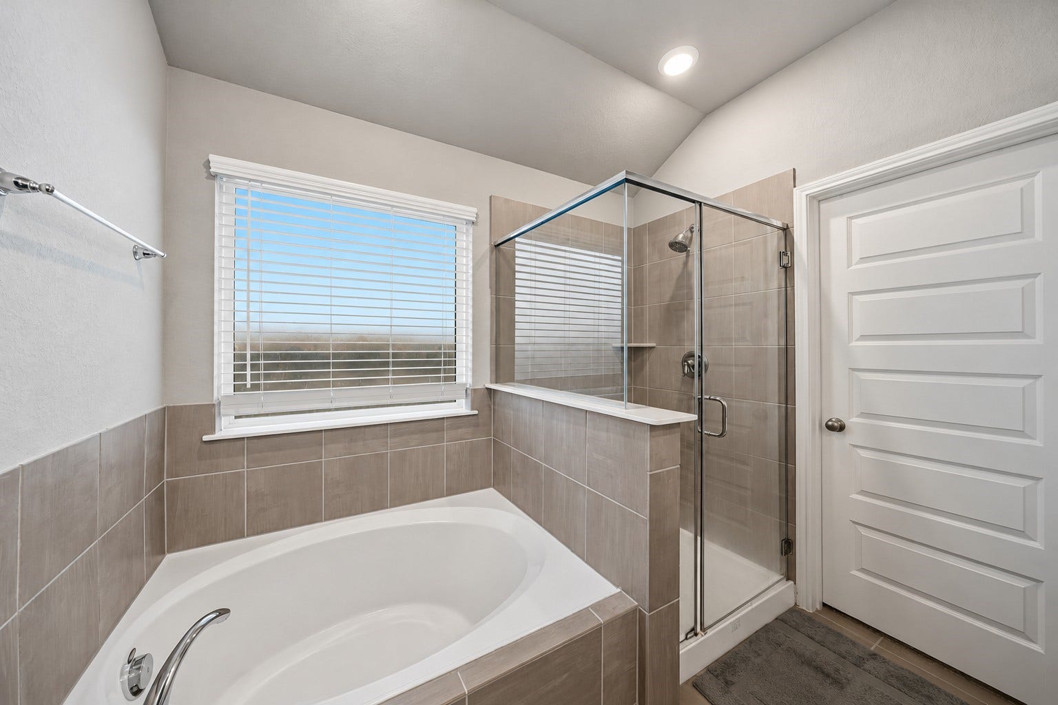 903 Pismo Lane, Rosharon, TX 77583 Virtually Enhanced: This spacious primary bathroom features a relaxing soaking tub beneath a large window with white trim and horizontal blinds, allowing natural light to brighten the space. A separate glass-enclosed walk-in shower with a chrome-framed door provides added convenience and functionality. Neutral-toned tile surrounds complement the walls, while the vaulted ceiling enhances the open, airy atmosphere.
