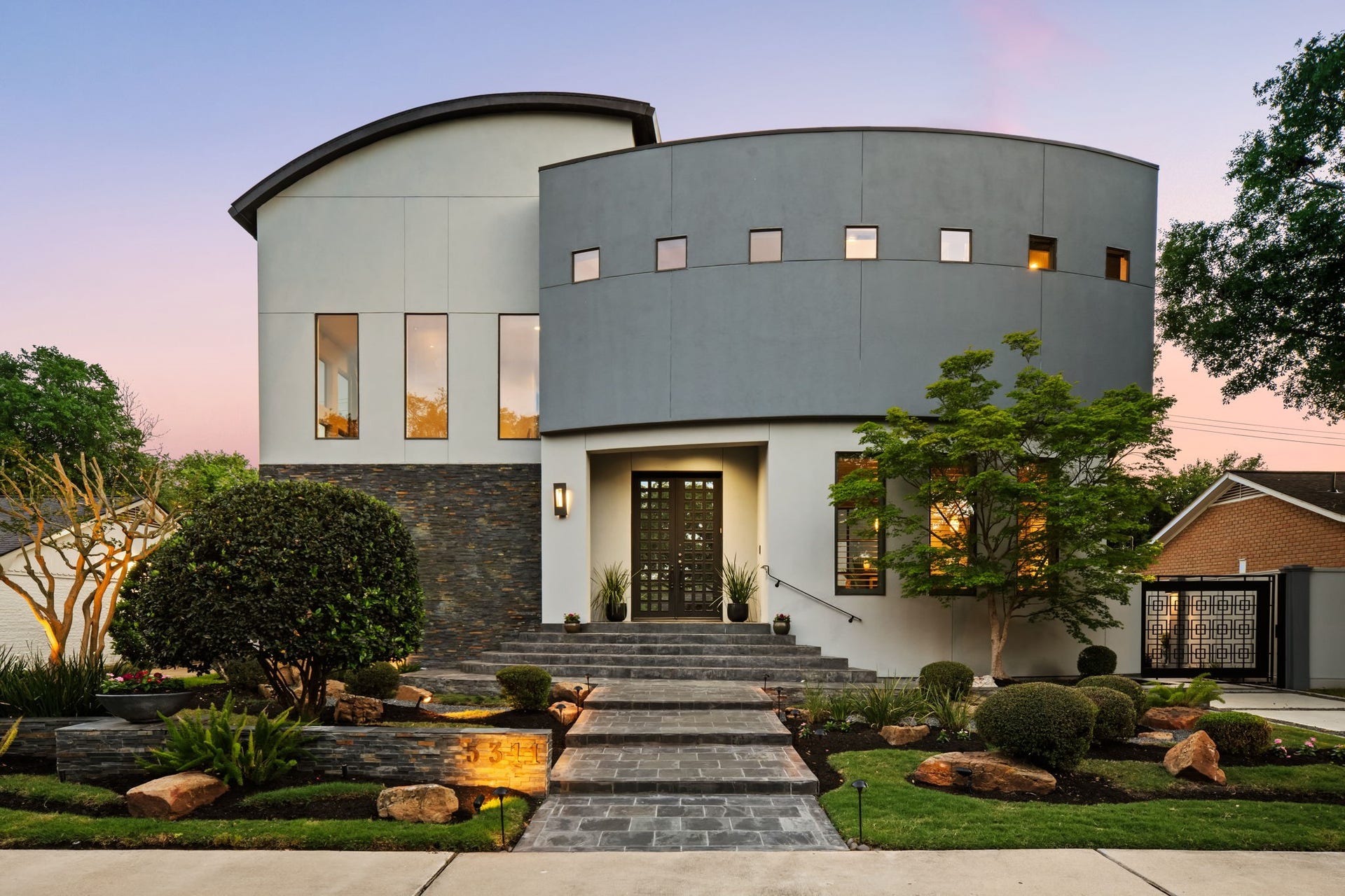 5311 Queensloch Drive, Houston, TX 77096 This contemporary two-story home boasts a distinctive curved design. The beautifully landscaped front yard features a charming pathway that leads to a sleek front door, enhanced by elegant outdoor lighting.