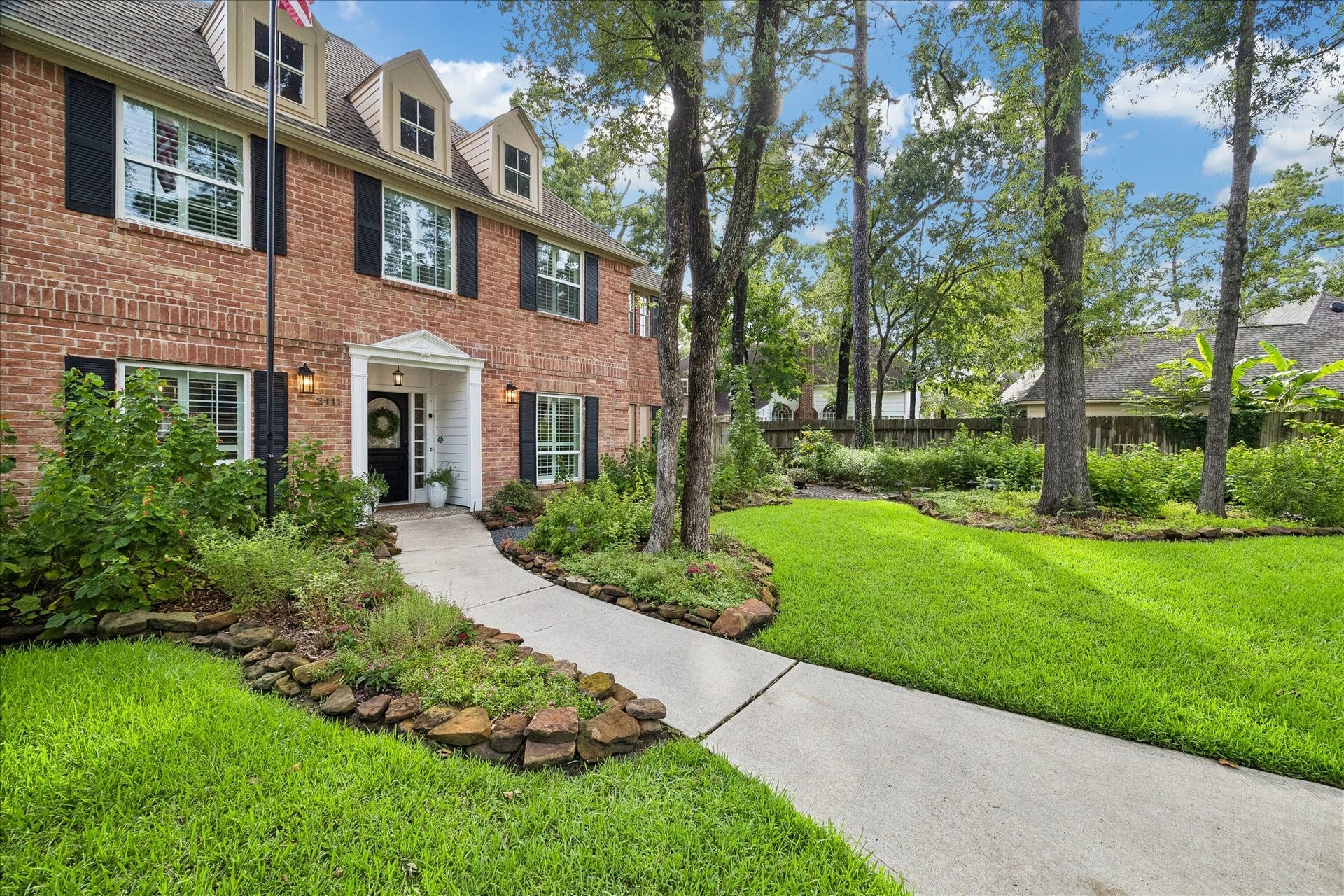 2411 Kings Lodge Drive, Kingwood, TX 77345 Charming two-story brick home with classic black shutters, nestled among mature trees. A welcoming front path leads through lush, landscaped gardens to the inviting entrance.