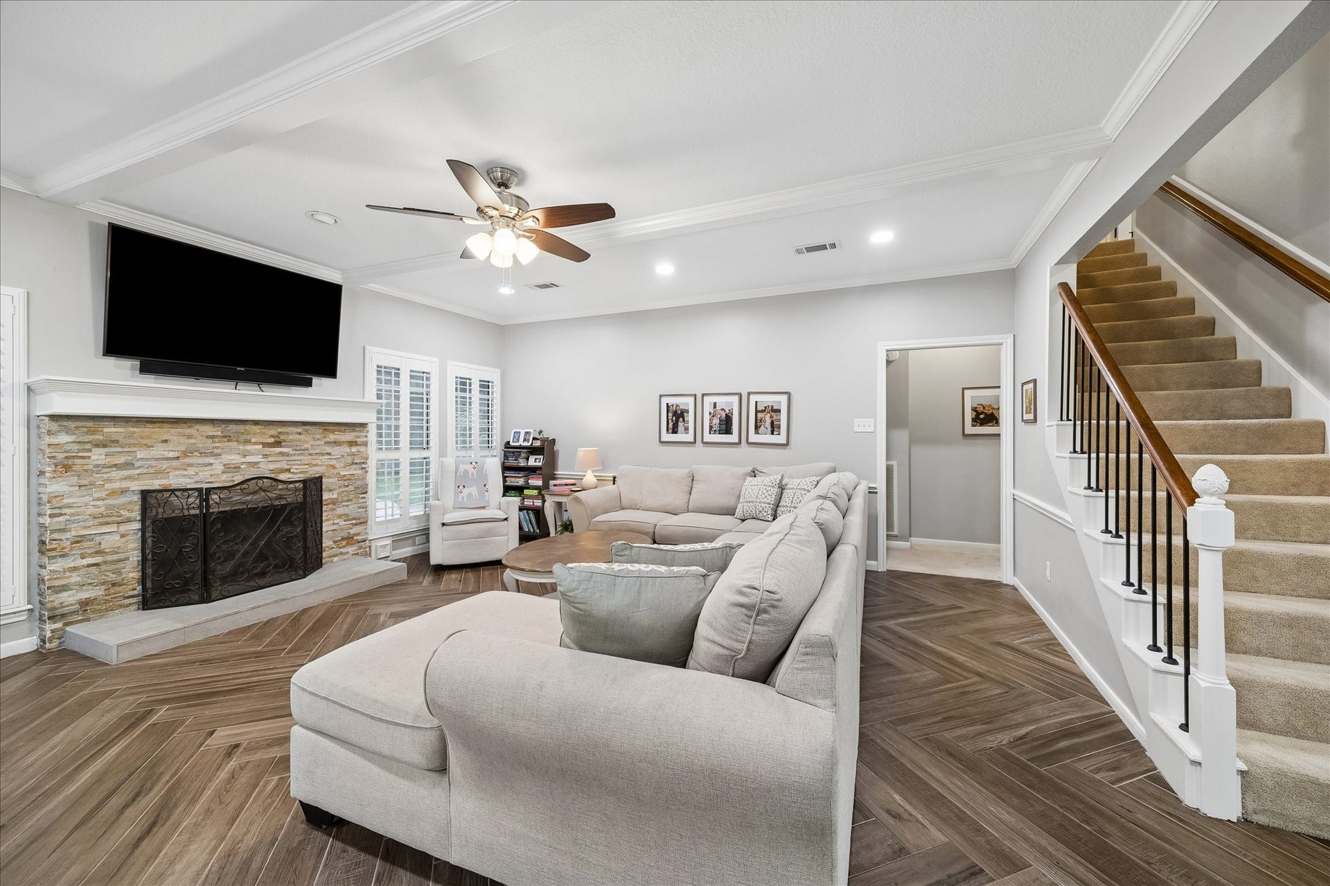 2411 Kings Lodge Drive, Kingwood, TX 77345 Spacious living room featuring a cozy stone fireplace, elegant herringbone wood flooring, and a stylish ceiling fan. The room is complemented by a comfortable sectional sofa and a sleek staircase leading to the upper level.