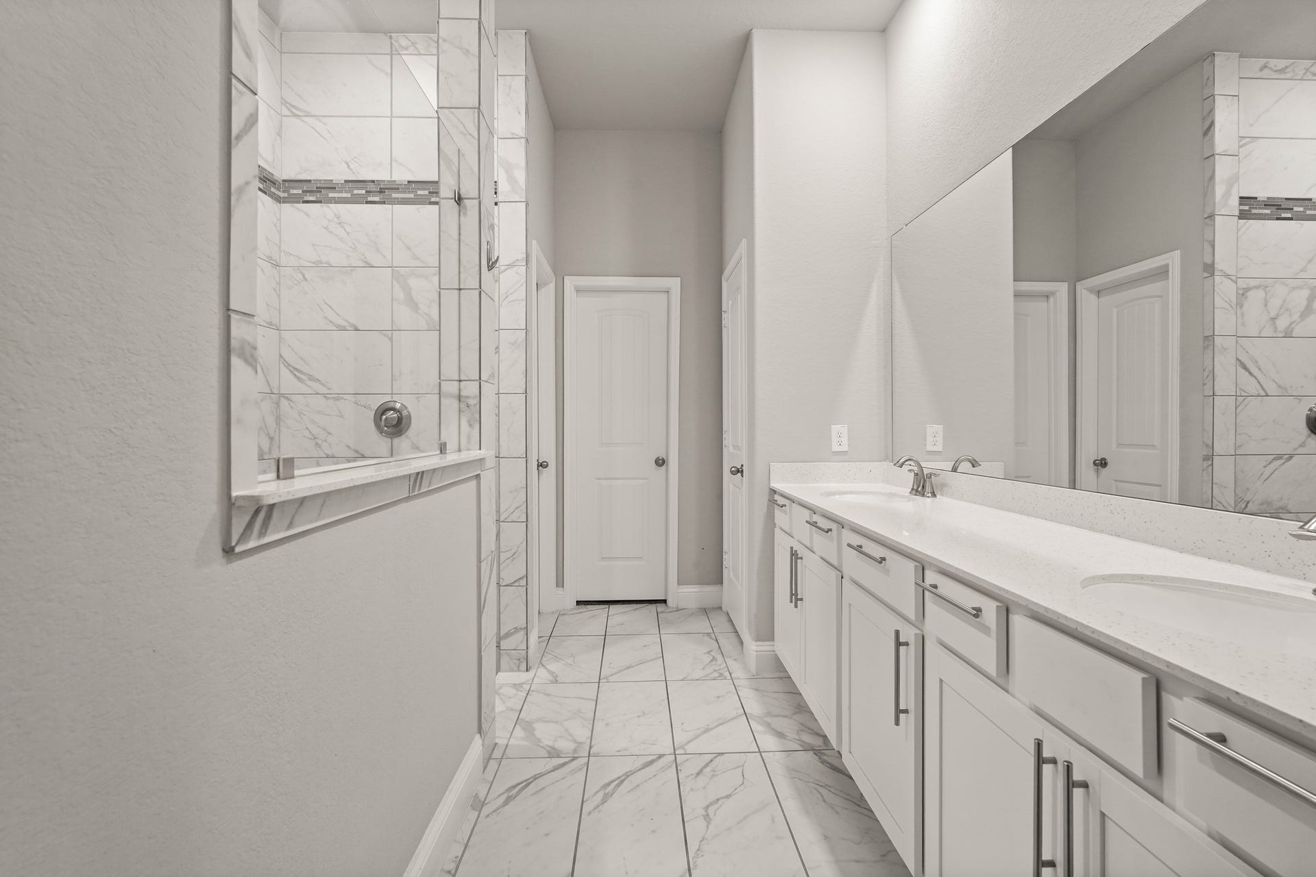 1710 Forest Mist Drive, Missouri City, TX 77459 This spacious main bathroom features a long, functional layout with clean finishes. On the right, a wide double vanity offers plenty of storage with white cabinets, long silver handles, and a bright white countertop with dual sinks under a massive wall mirror. On the left, the walk-in shower is styled with large-format marble-look wall tile and a dark mosaic accent strip.