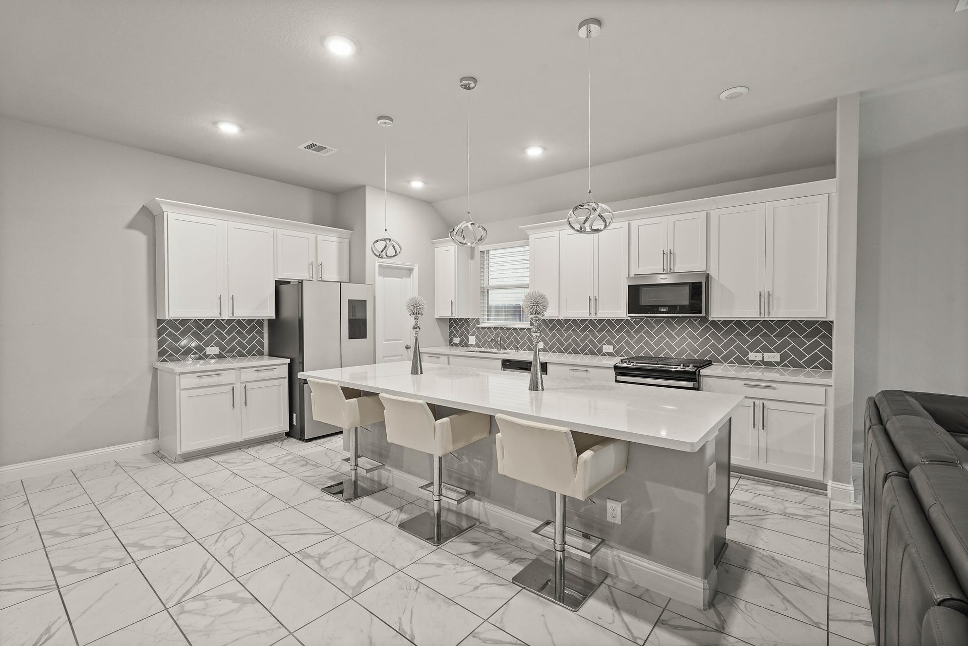 1710 Forest Mist Drive, Missouri City, TX 77459 This bright, contemporary kitchen features an expansive island with seating and a seamless white polished countertop. The design includes timeless white cabinetry with sleek silver hardware, a striking grey herringbone tile backsplash, and matching solid surfaces. Modern spherical pendant lights, recessed ceiling lights, and elegant marble-look tile flooring beautifully complete the space.
