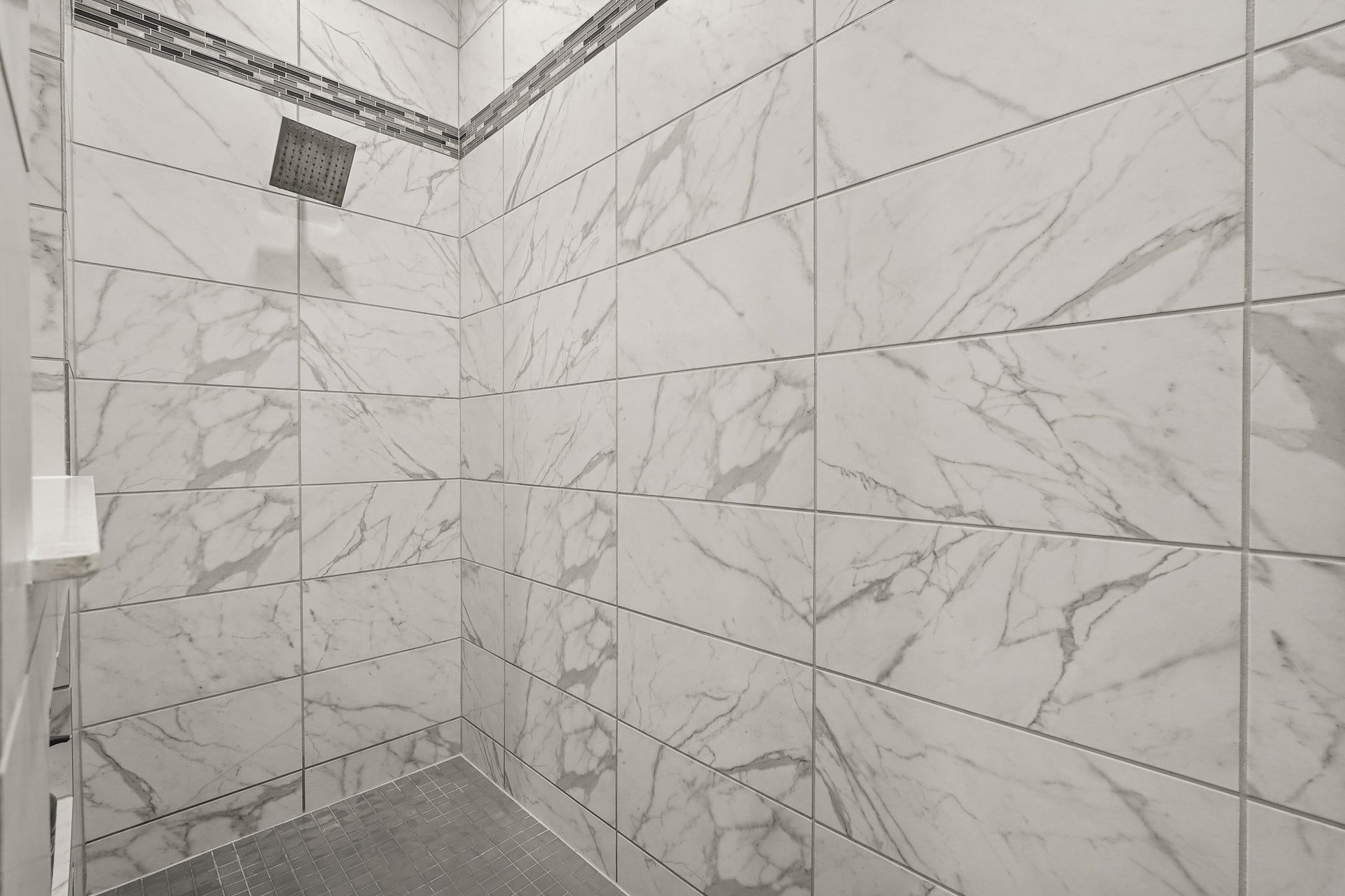 1710 Forest Mist Drive, Missouri City, TX 77459 This detailed look inside the custom walk-in shower, highlights its clean and modern design. The spacious enclosure is fully lined with large-format wall tiles featuring a bright white and grey marble pattern. A stylish horizontal mosaic tile accent strip runs along the upper perimeter of the walls.