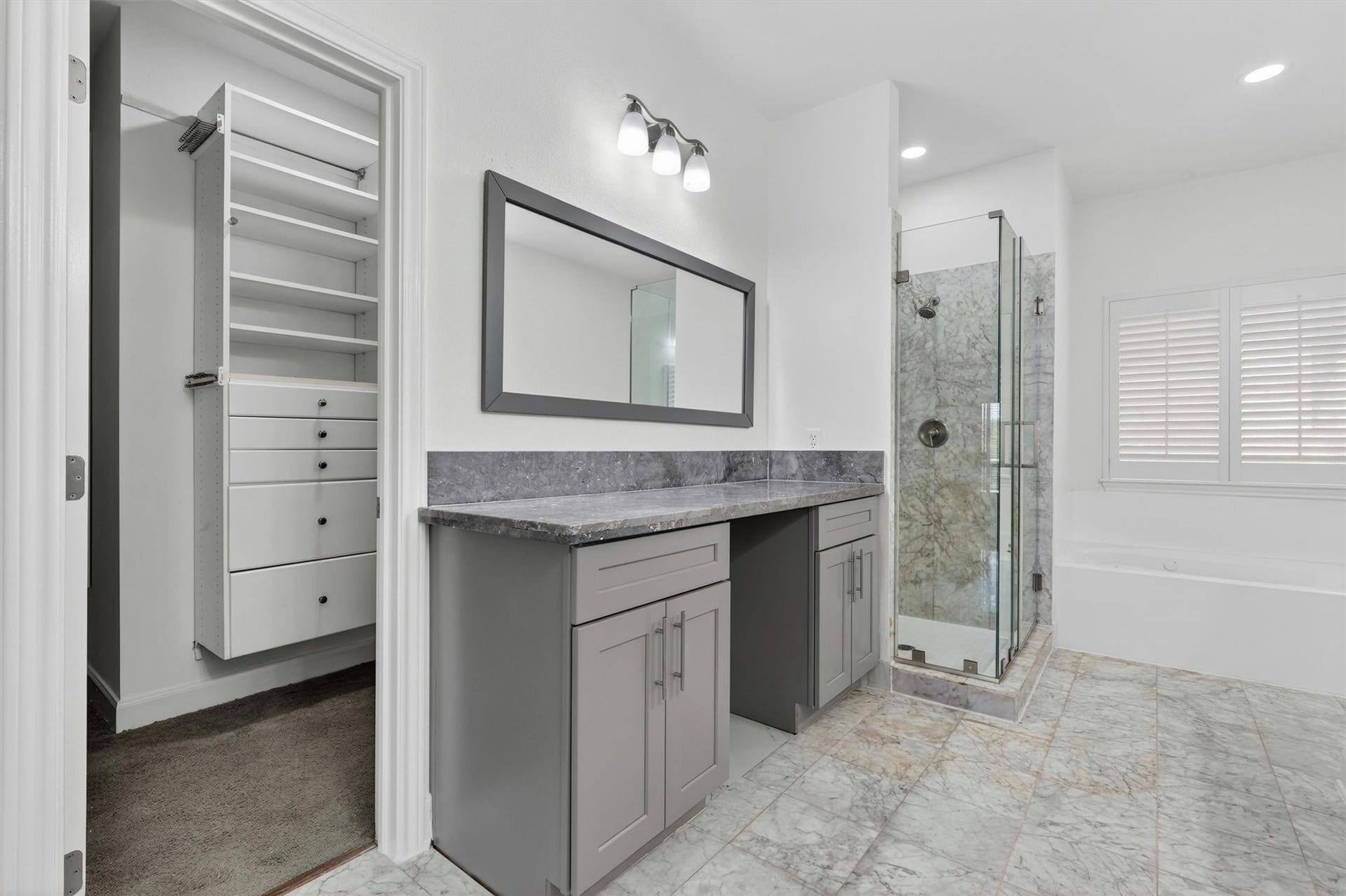 4720 Post Oak Timber Drive, #23, Houston, TX 77056 Luxurious primary bathroom featuring marble-look tile flooring, oversized vanity with granite countertops, framed mirror, and designer lighting. Spa-inspired walk-in shower with frameless glass enclosure, soaking tub, plantation shutters, and custom walk-in closet with built-in shelving complete this elegant retreat.
