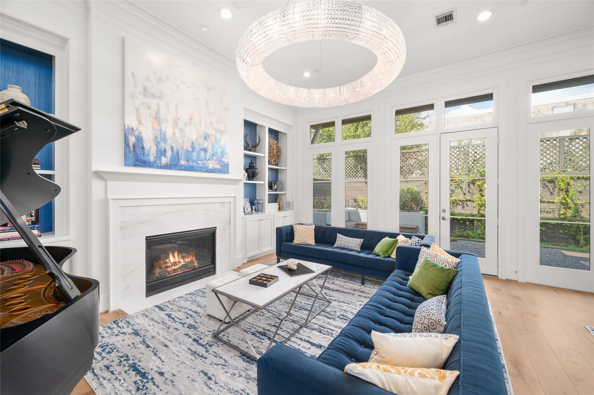 7554 Little Caprese Lane, Houston, TX 77055 Sunlit living room with a marble-clad fireplace, dramatic circular chandelier, and a wall of windows and French doors opening to the courtyard.