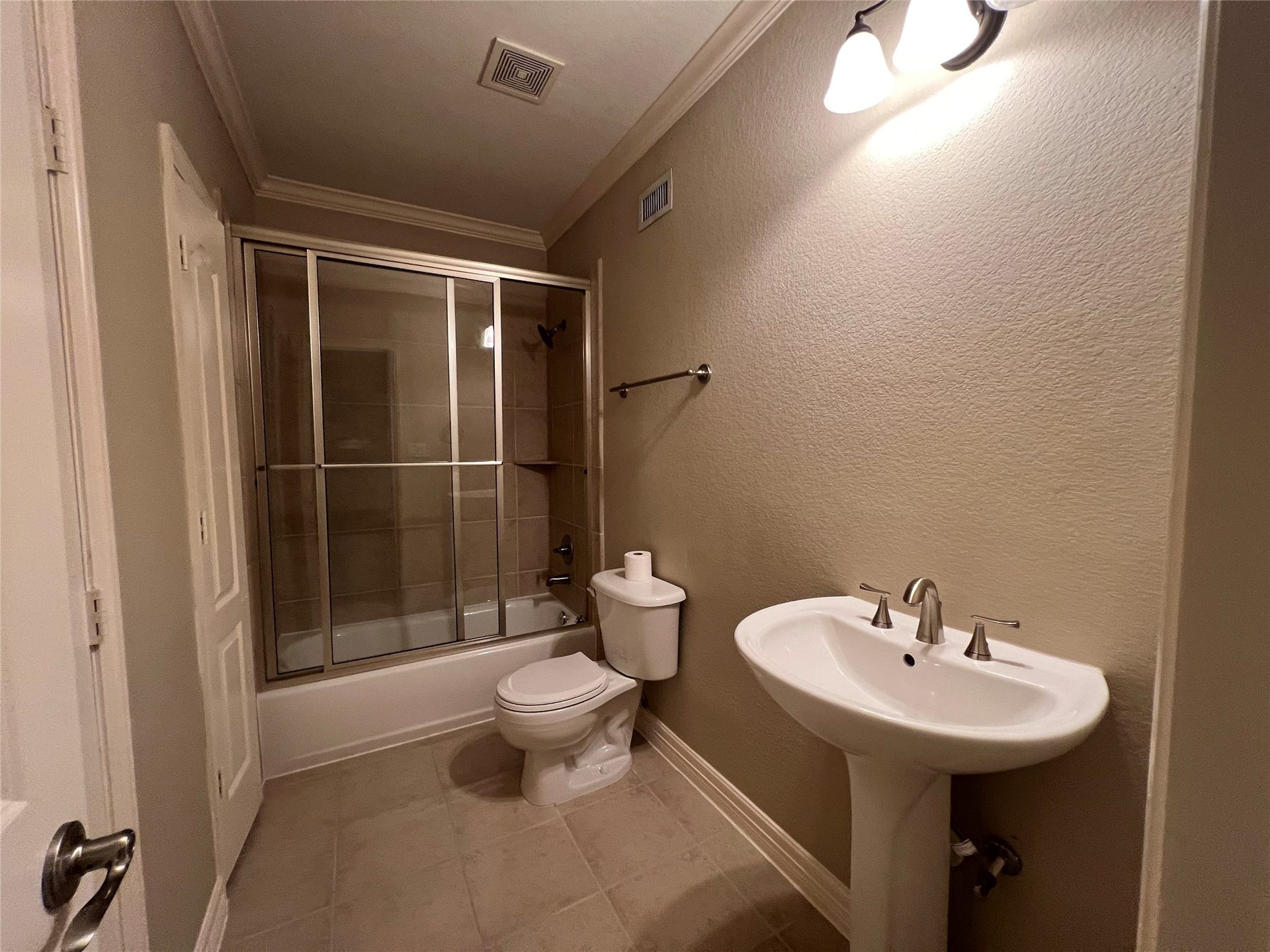 1625 Cohn Street, Houston, TX 77007 This bathroom features a bathtub with sliding glass doors, a pedestal sink, and a toilet. The neutral tile flooring and walls create a clean, modern look, complemented by overhead lighting.