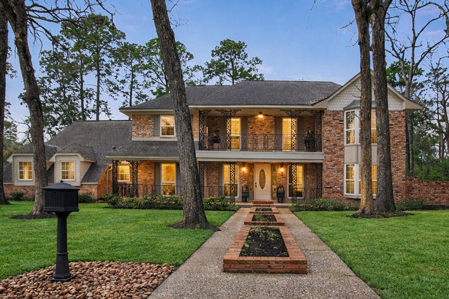 112 Kirkwood Lane, Conroe, TX 77304 Stately two-story brick home with elegant wrought iron accents, surrounded by mature trees and lush landscaping, offers charming curb appeal and a welcoming pathway.