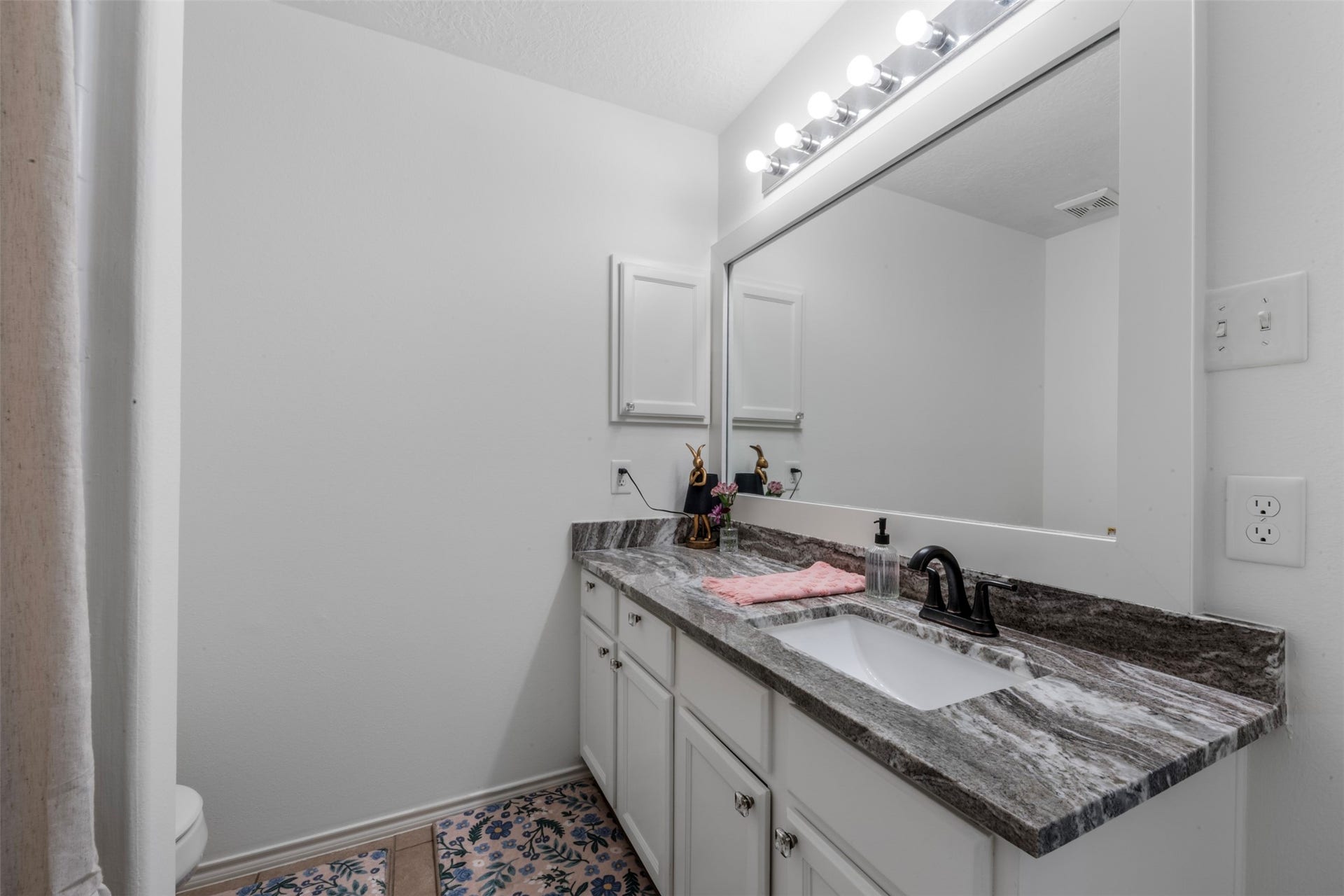 9750 Gulfstream Drive, Conroe, TX 77303 This bathroom features a sleek granite countertop with an undermount sink, complemented by a large mirror with bright lighting. The vanity offers ample storage with white cabinetry, and the space is accented by a patterned tile floor.