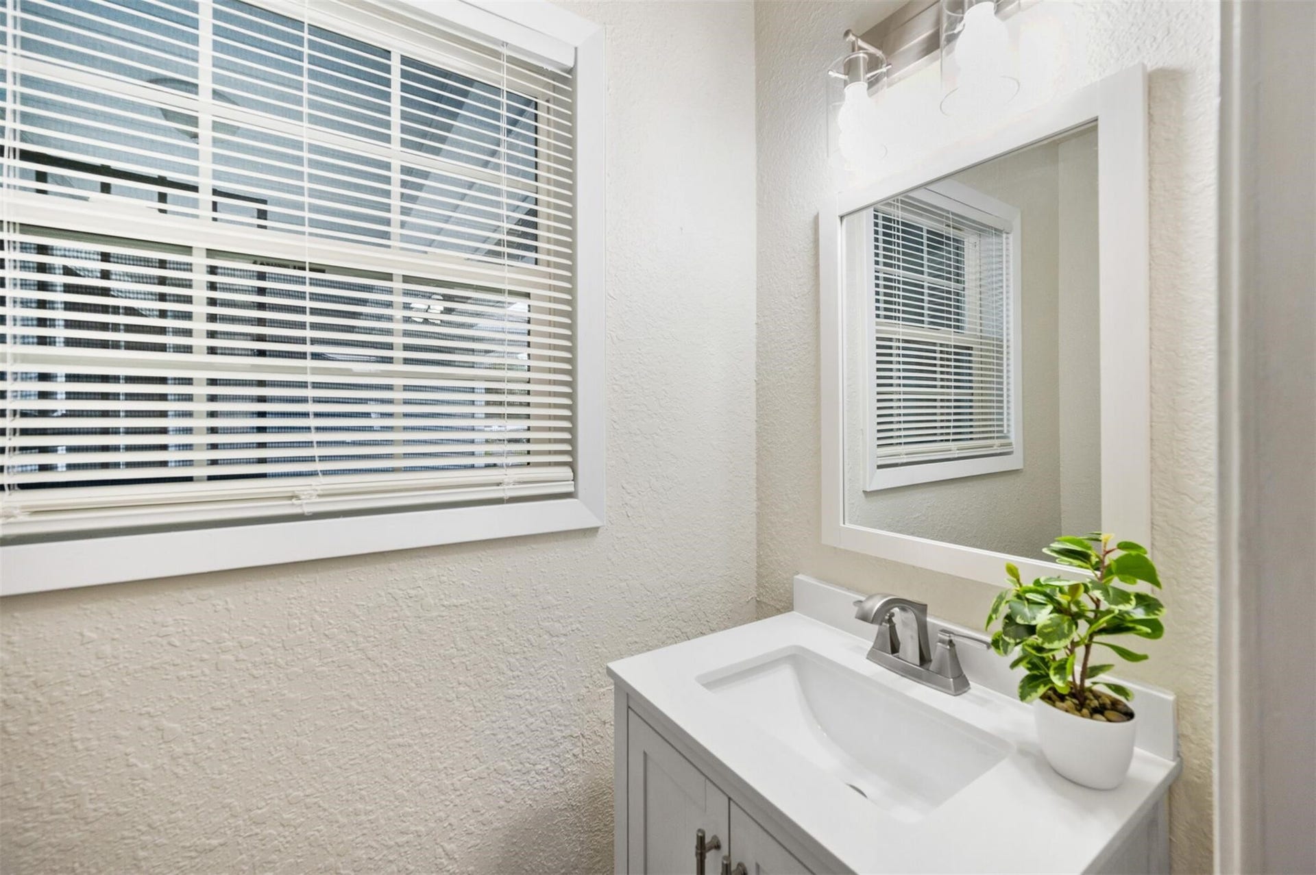 Bright and inviting powder room featuring a modern vanity with a sleek mirror, complemented by a touch of greenery and ample natural light from the window.