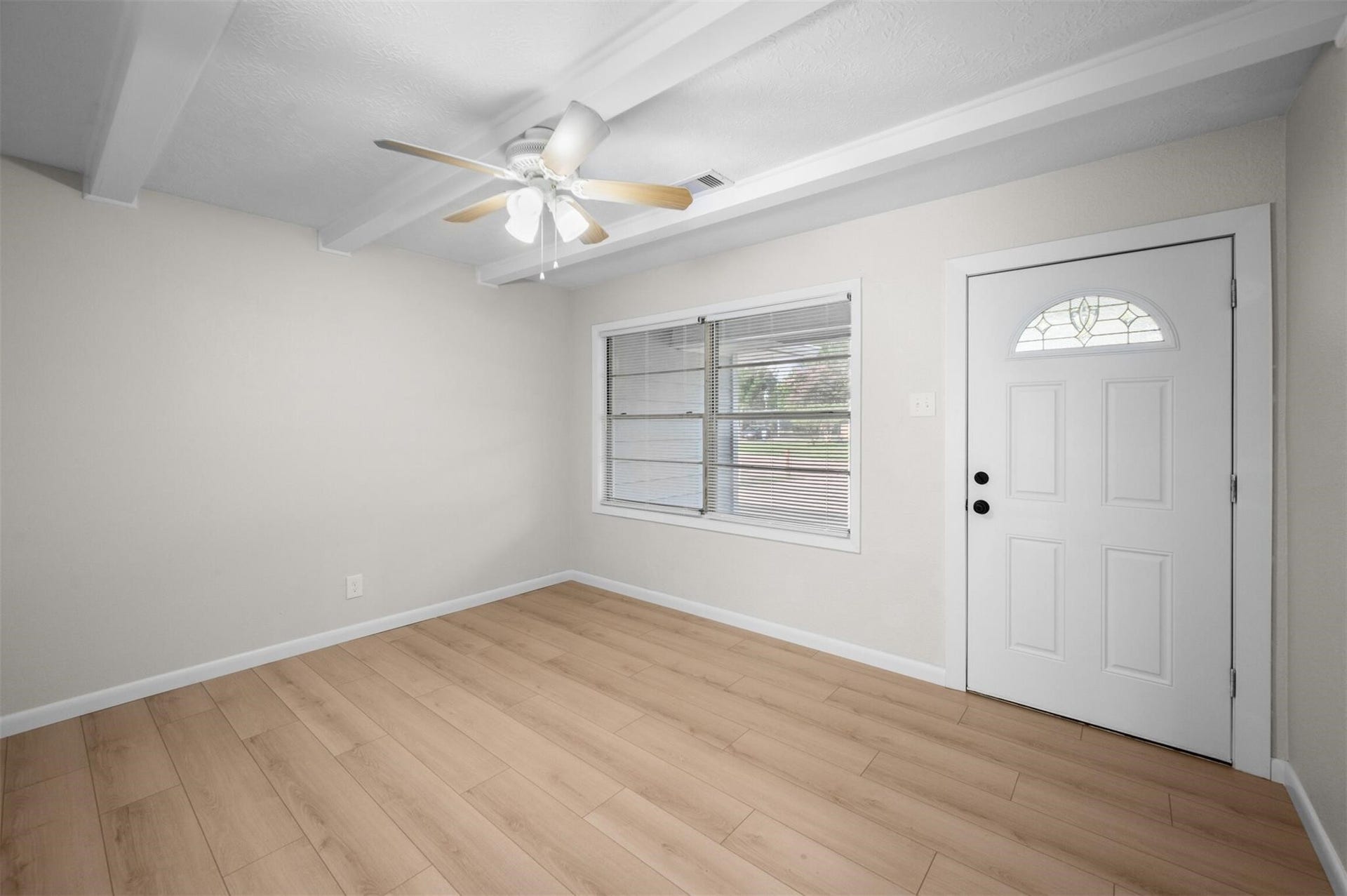 Bright and inviting room featuring wood-look flooring, a ceiling fan with light, and ample natural light from a large window, complemented by a classic white front door.