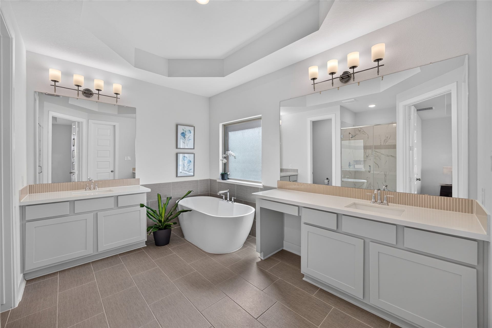 7311 Winding Aster Way, Katy, TX 77493 The primary bathroom features two separate vanity areas with gray cabinetry, broad mirrors and decorative lighting. A freestanding soaking tub sits beneath the window between the vanities, creating a focal point. A tiered ceiling adds architectural detail, while cabinetry and counter space support everyday routines.