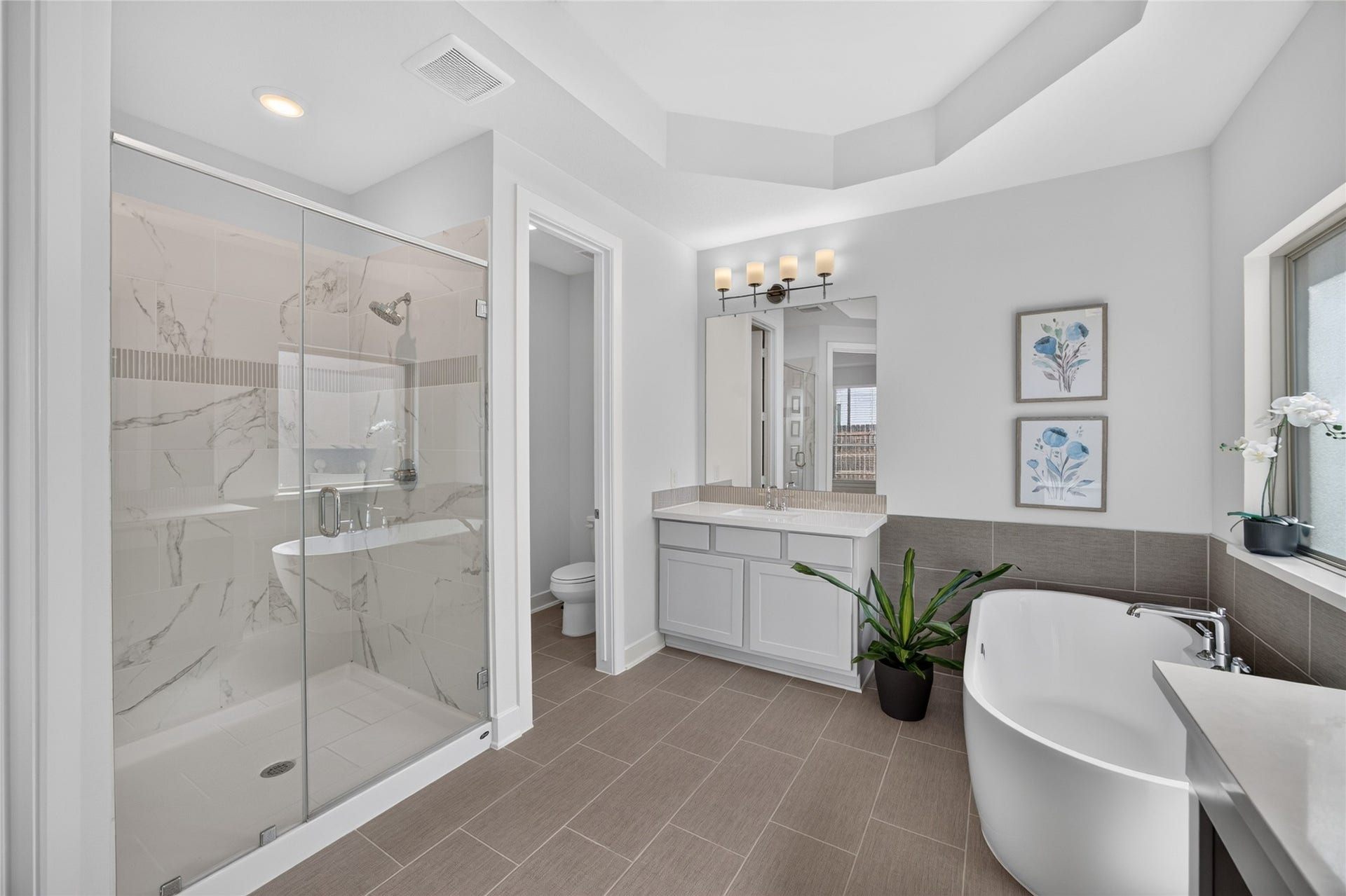 7311 Winding Aster Way, Katy, TX 77493 A glass-enclosed shower showcases large-format patterned tile and a coordinating decorative accent band. The adjacent freestanding tub provides a separate bathing option, while the nearby vanity offers storage and counter space. Natural light and neutral finishes give the room a bright, cohesive appearance.